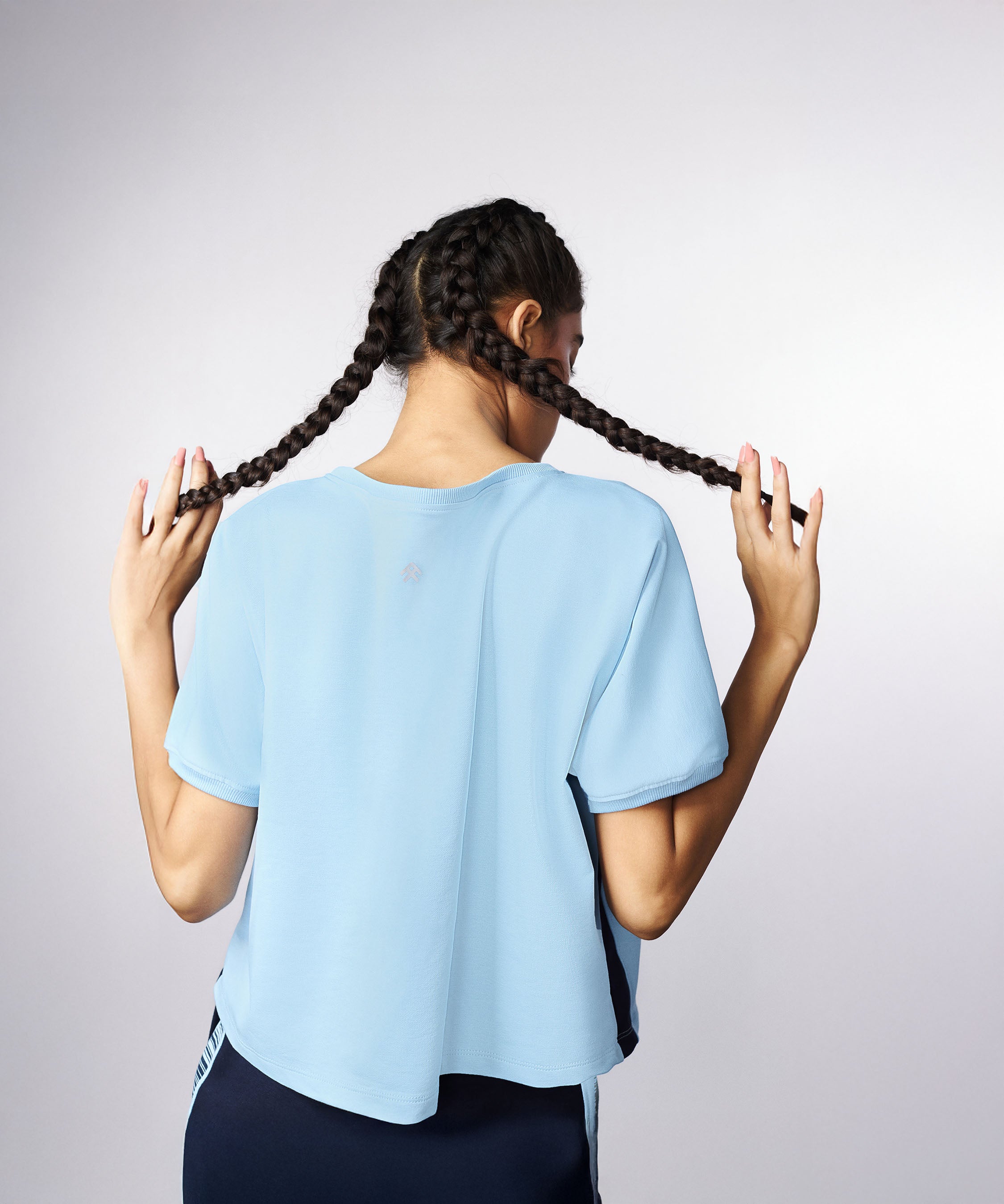 A woman with two braids, facing away and holding her hair, is wearing the Tuna Active Blue Tee and dark pants.