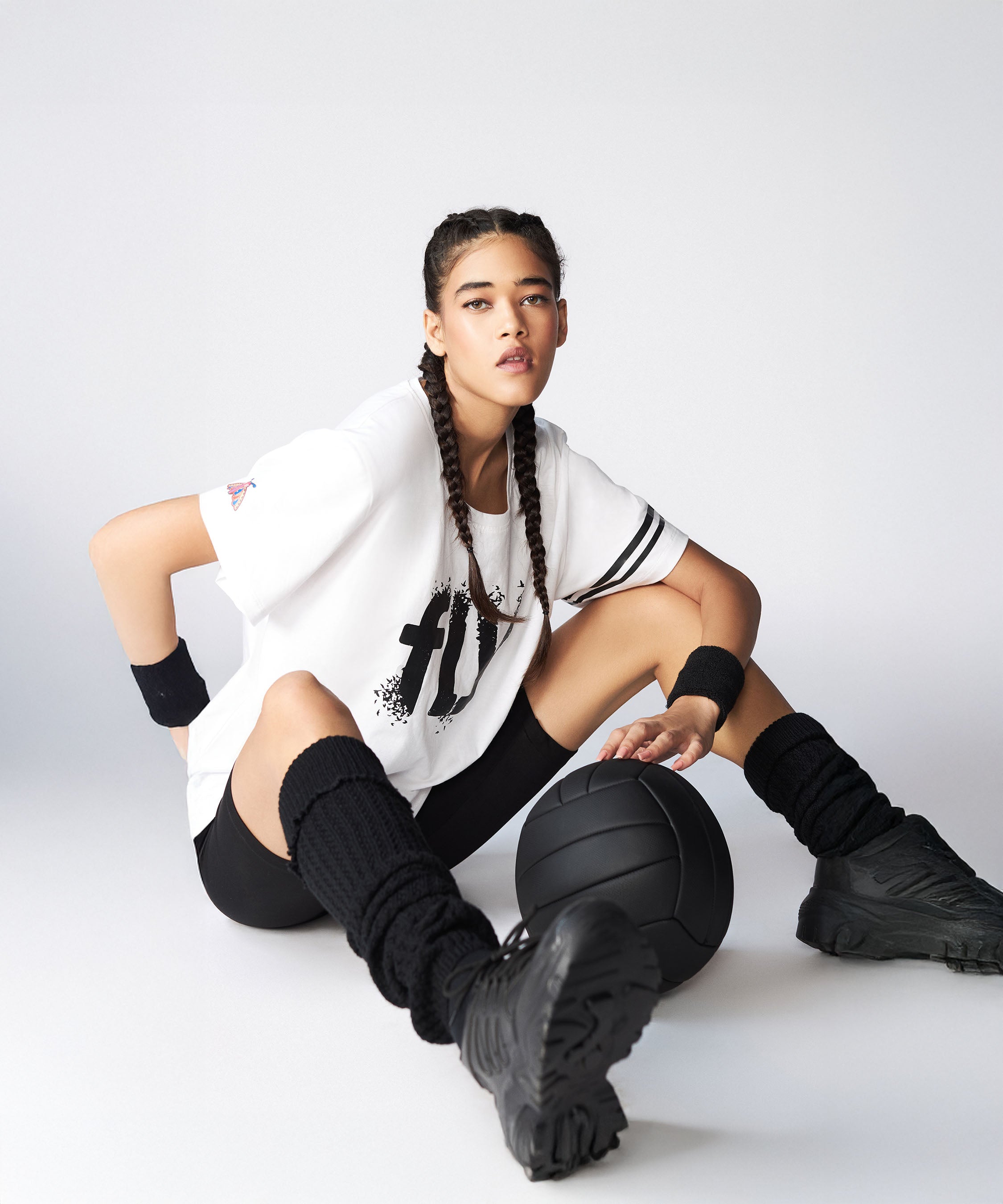 A woman in sportswear with braids sits on the floor wearing the Oversize Fly Tee, confidently holding a basketball and looking at the camera.