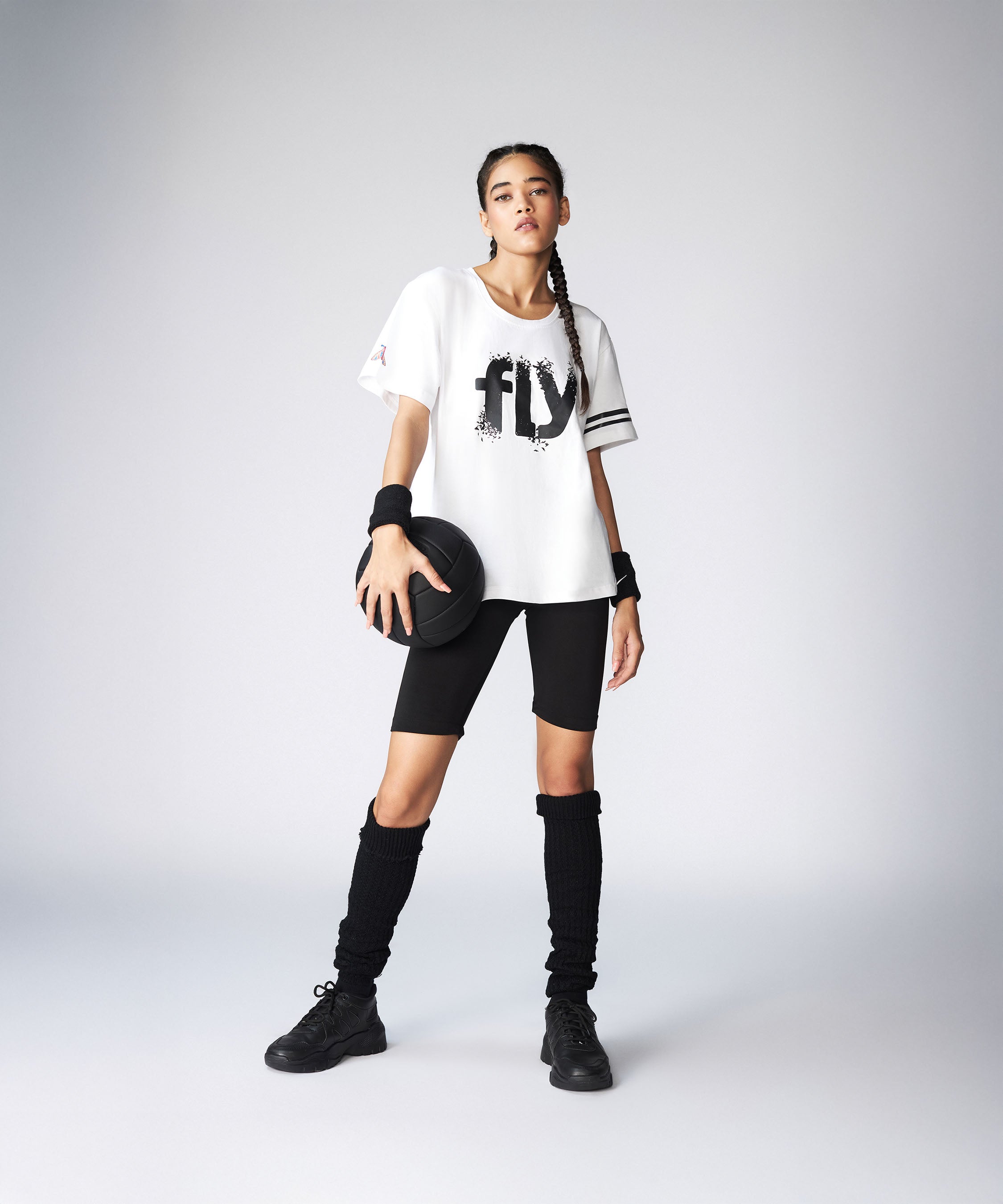 A young woman in a sporty outfit holds a basketball, wearing the Oversize Fly Tee with bold "FLY" print, black shorts, and long socks. The 100% cotton Oversize Fly Tee offers comfort both on and off the court.