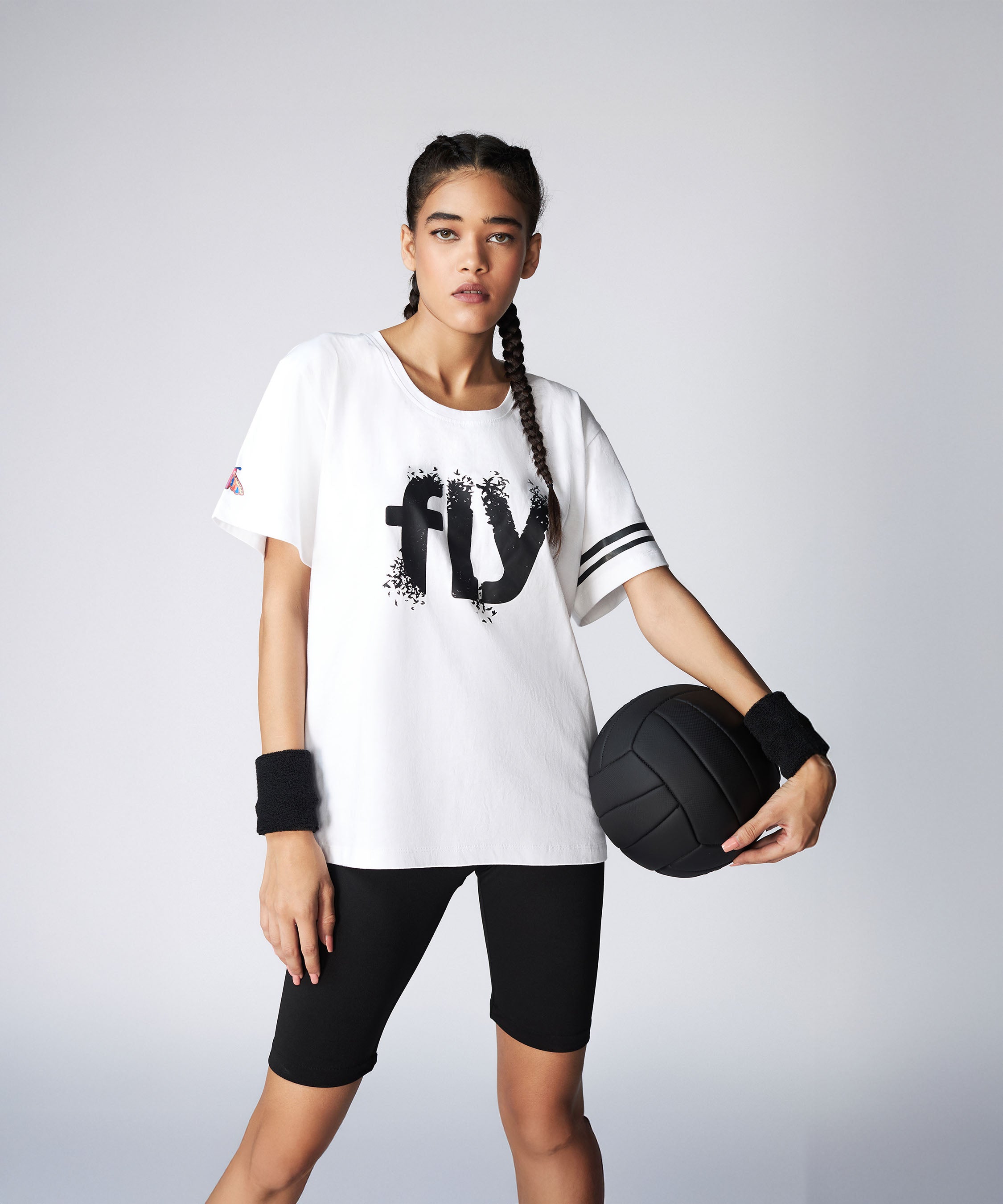 Woman in sporty outfit holding a black ball, wearing the Oversize Fly Tee—a 100% cotton butterfly print T-shirt—paired with black shorts and wristbands.