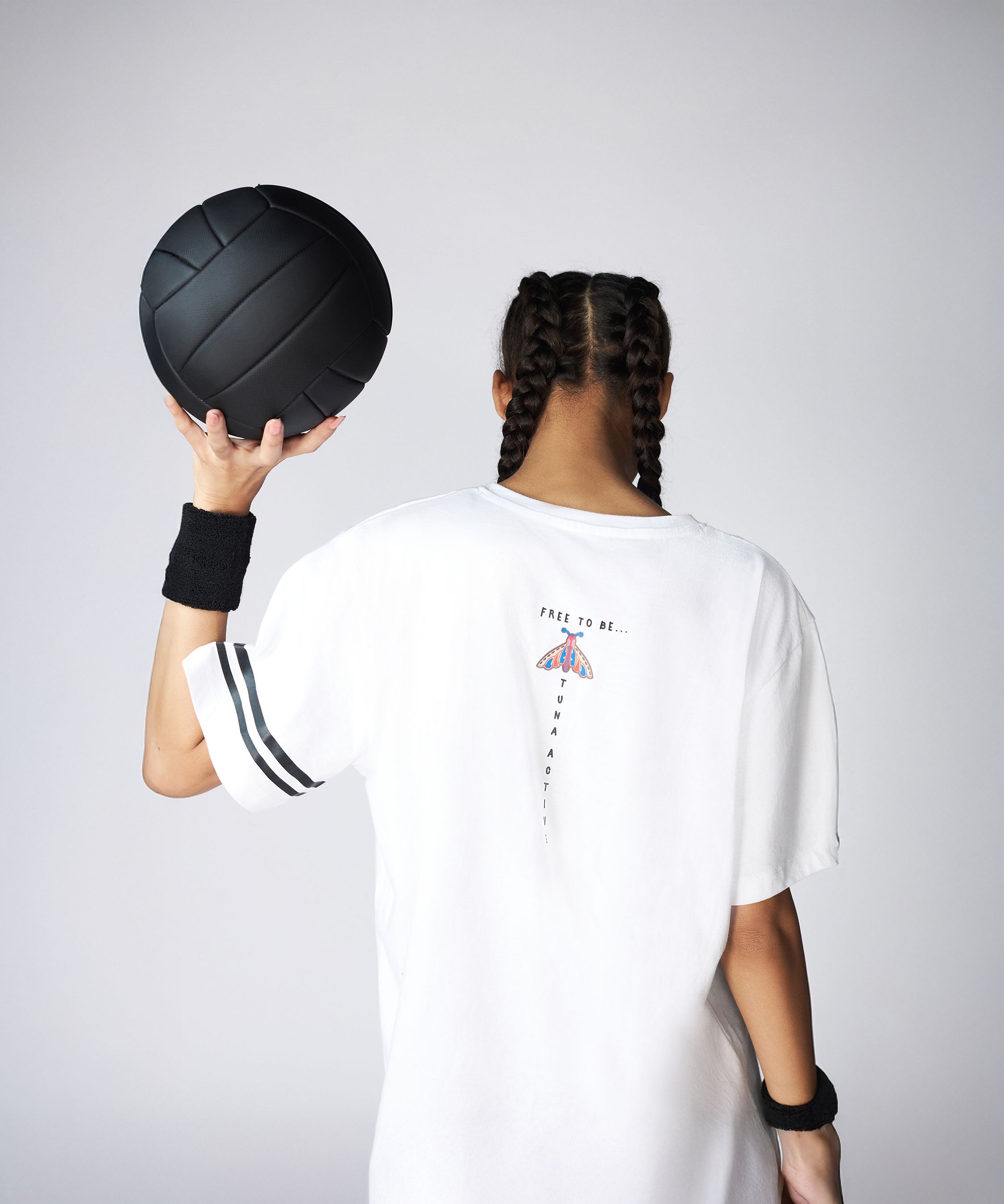 Person with braids, facing away, holding a black volleyball. Wearing the Oversize Fly Tee with a butterfly print and black wristbands.