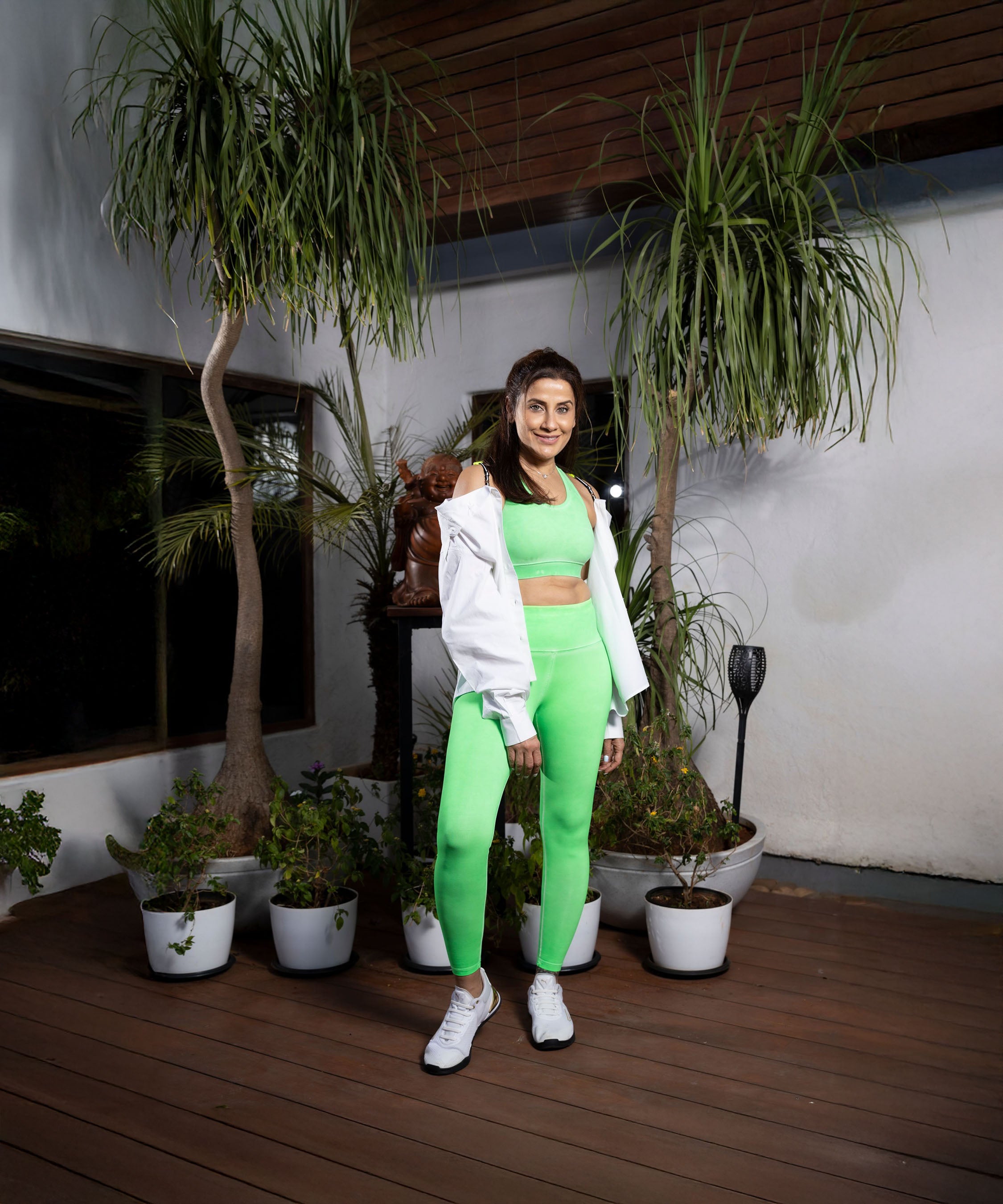 A woman wearing Neon Green Leggings and a white jacket stands on a wooden deck, with vibrant potted plants behind her.