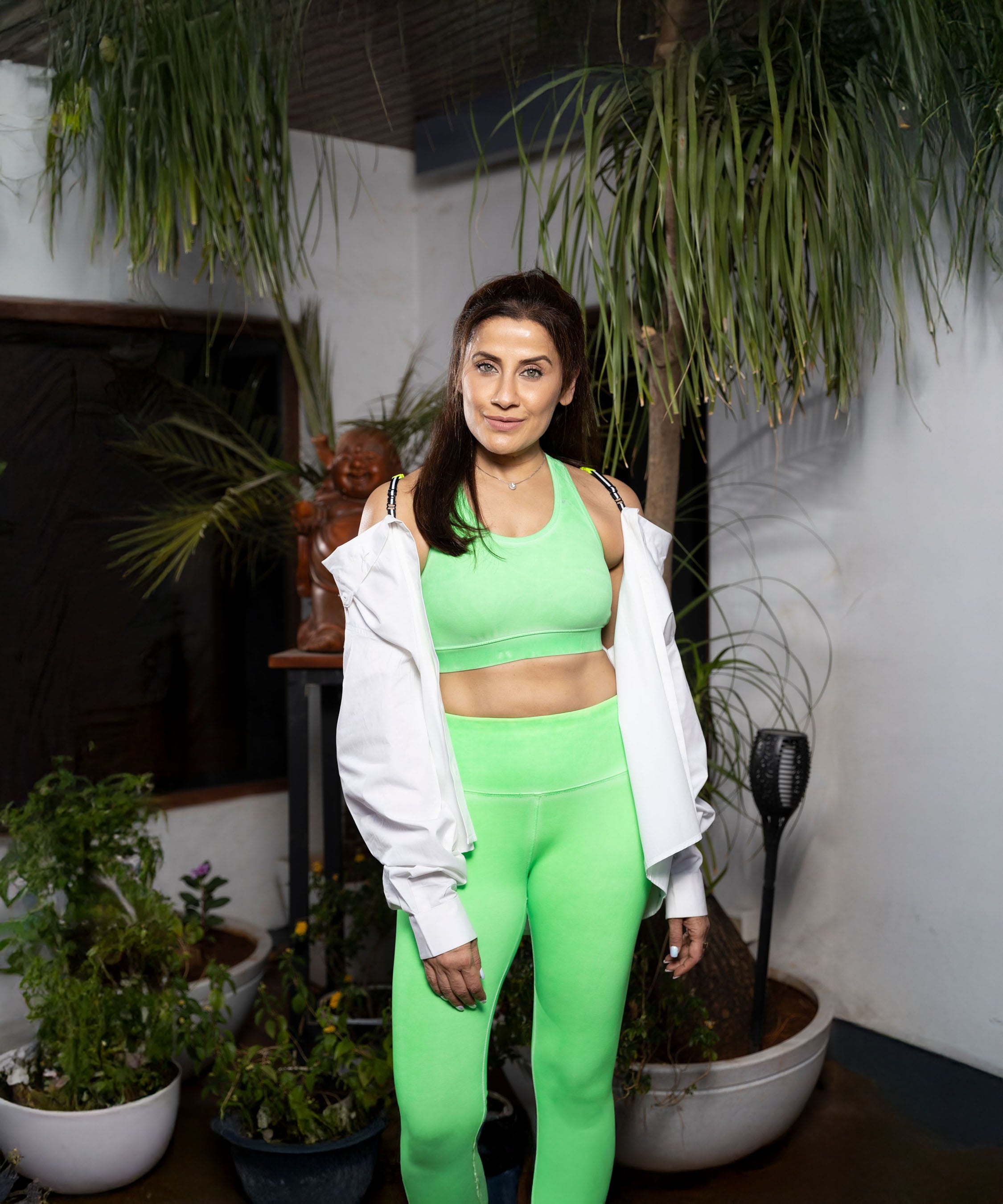 A woman wearing Neon Green Leggings and a white jacket stands by potted plants indoors, looking at the camera.