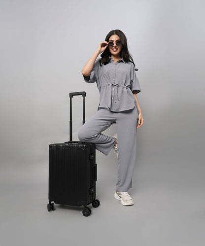 A woman poses confidently in a Grey Tie Up Top with Loose Pants Cord Set and sunglasses, one foot on a black suitcase, against a grey background.