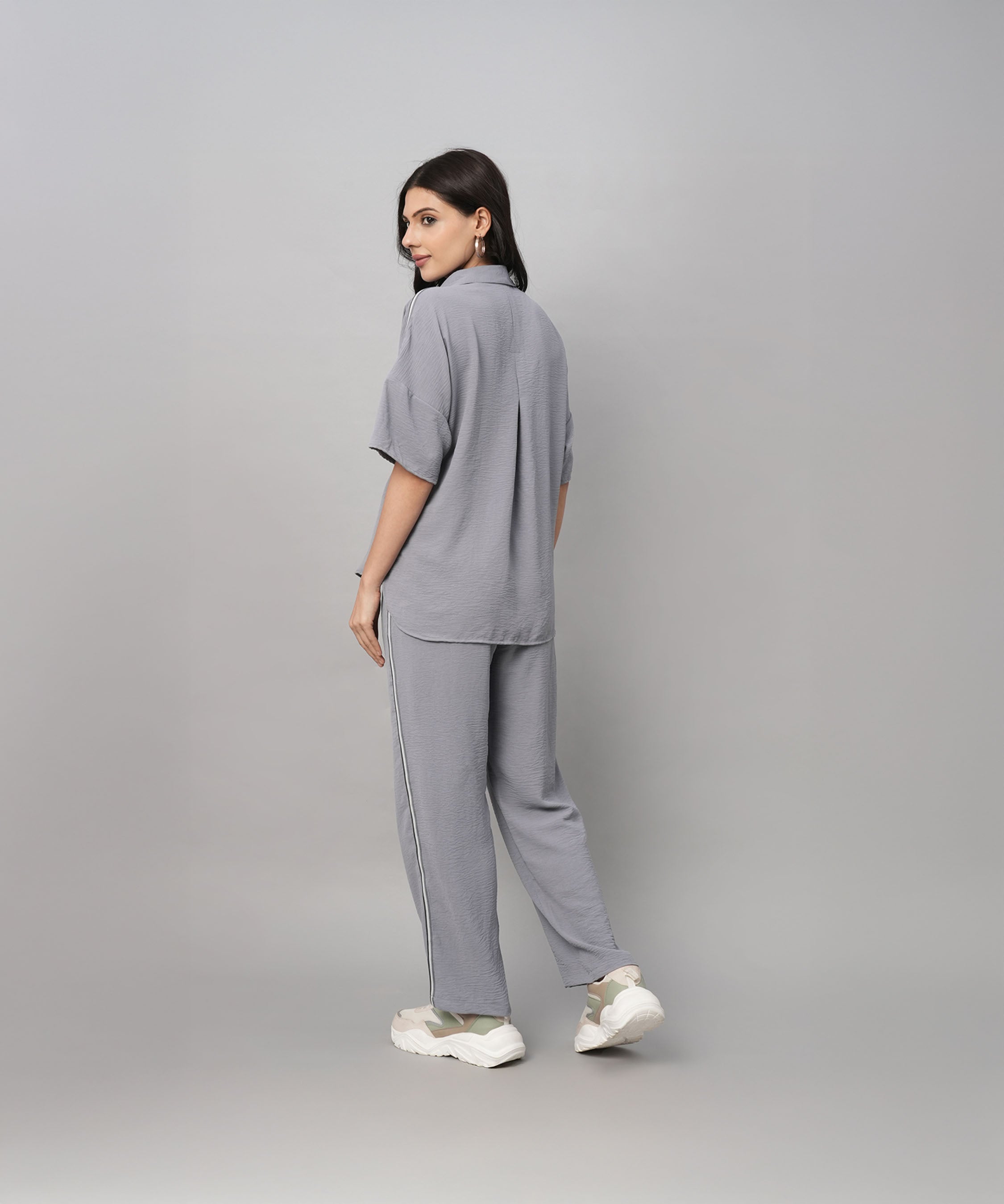 A woman in a Grey Tie Up Top with Loose Pants Cord Set and white sneakers stands slightly turned to the side against a grey background.