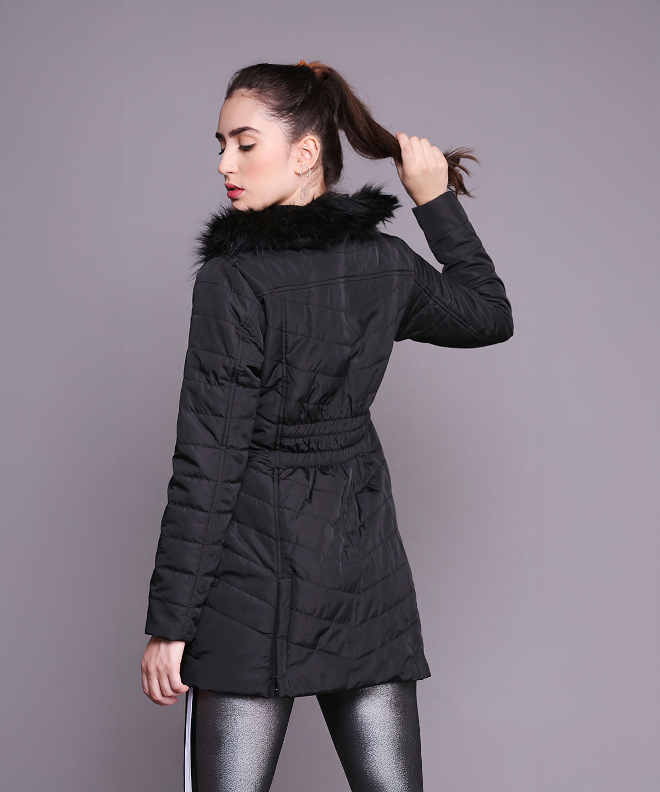 A woman faces away, holding her ponytail, wearing the Black Fur Collar Long Puffer Jacket With Button and Zip Details against a gray background.