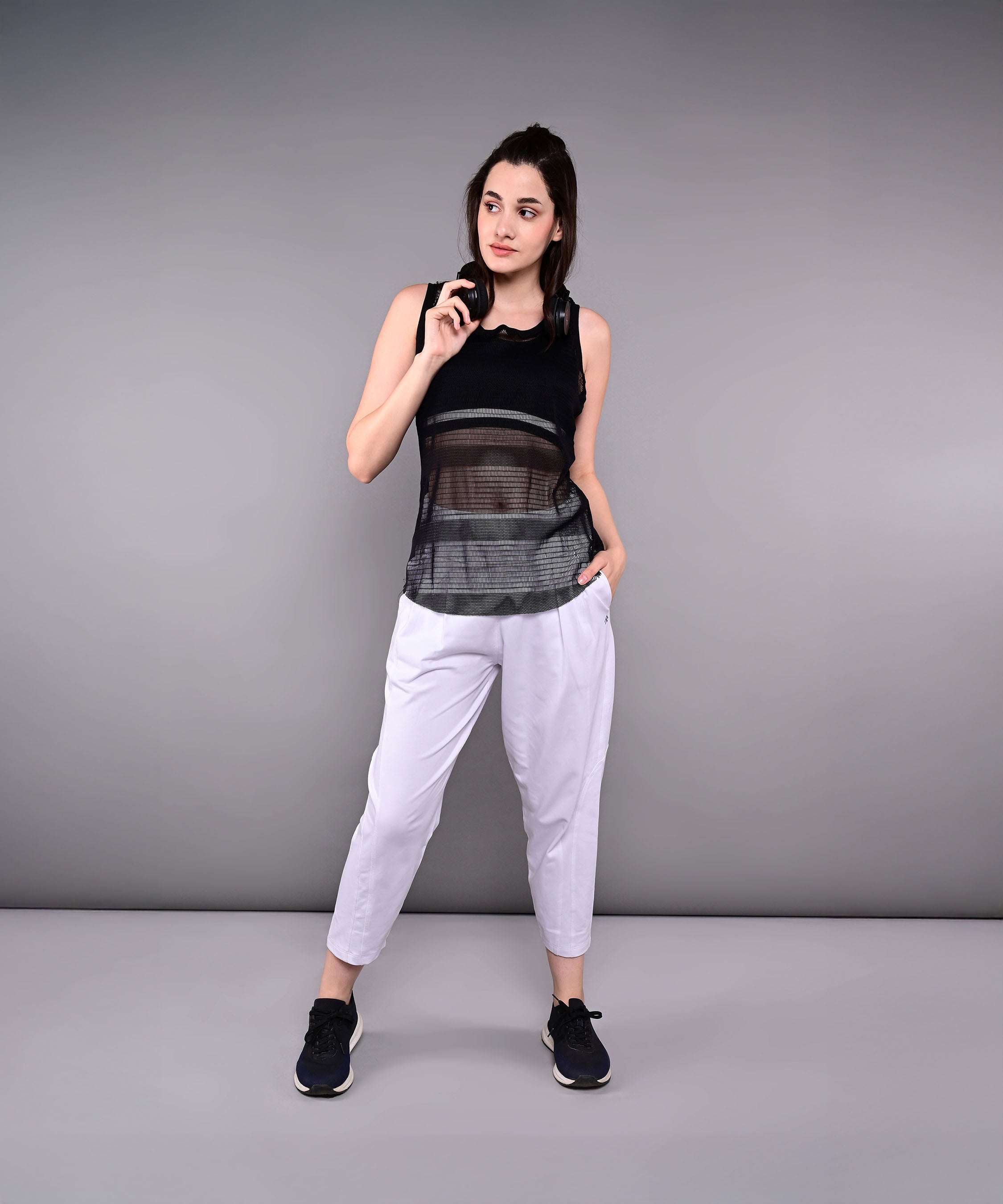 A woman in athletic wear poses against a gray background, holding sunglasses and with one hand in her pocket, showcasing the Pleated Yoga Pant With Smocked Waistband.