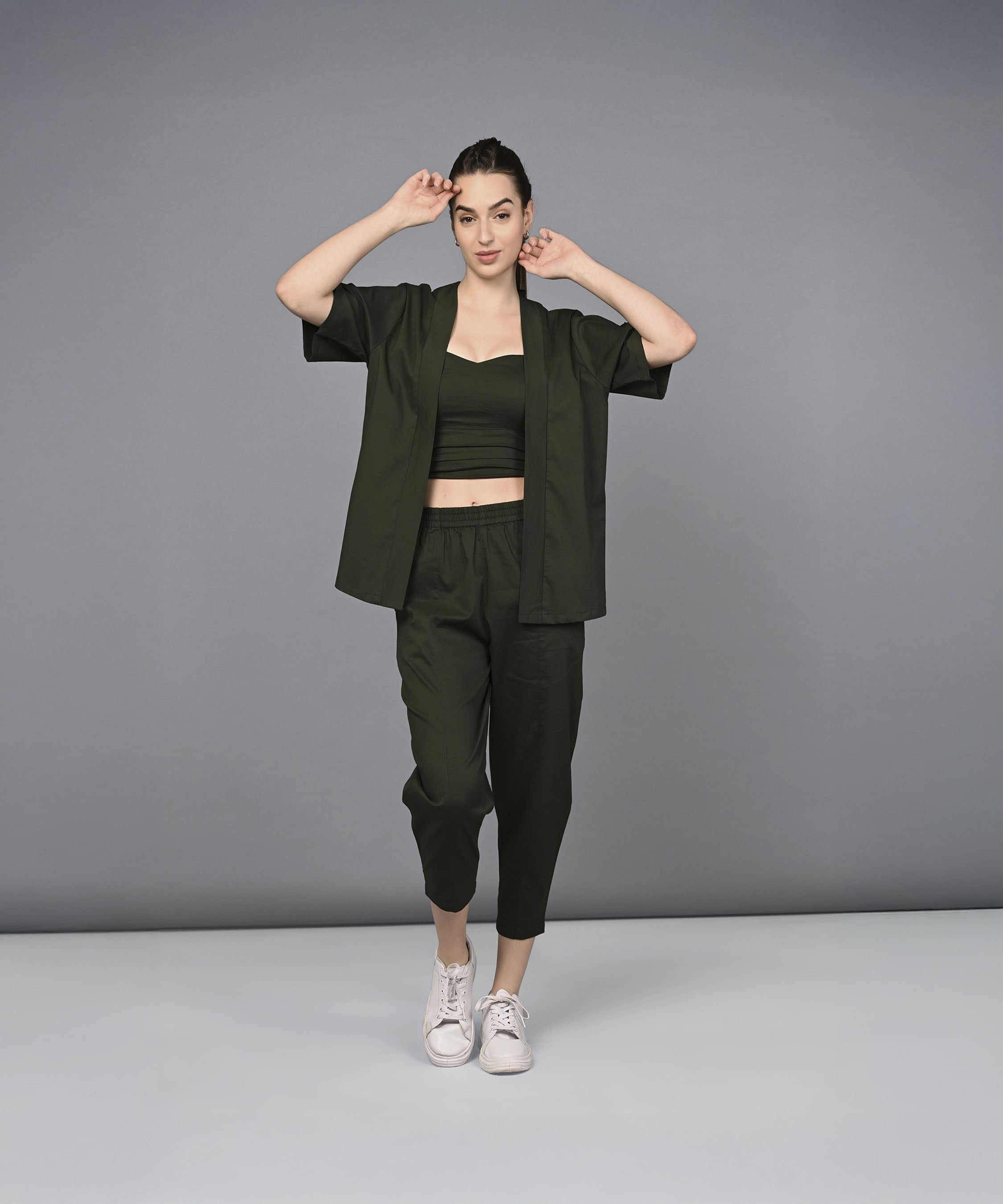 A woman models the Military Green Open Jacket & Yoga Pants with Bustiere set, standing against a gray background with hands near her head, styled with white sneakers.