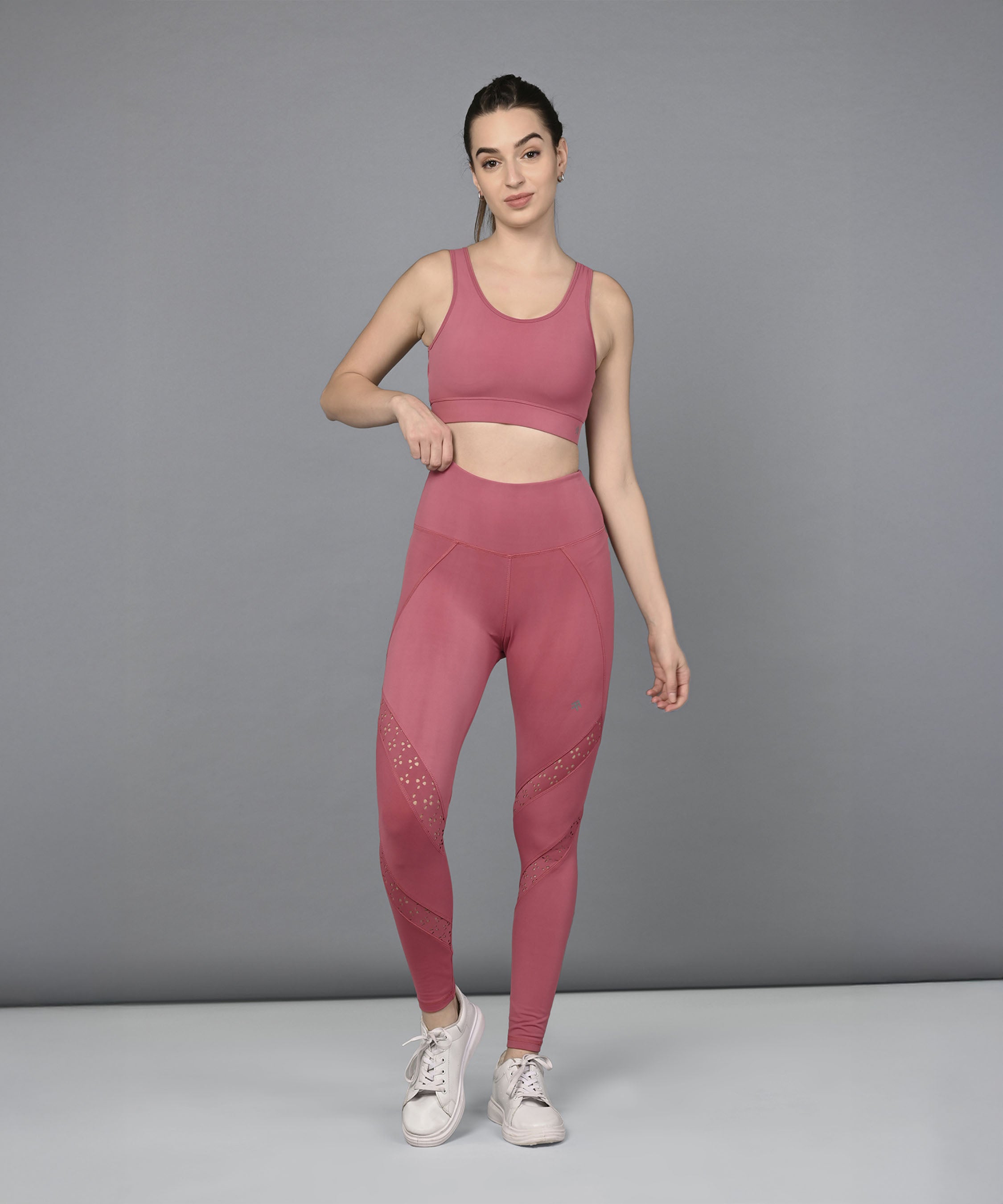 A woman wearing a Peony Pink Netted Top with a honeycomb self pattern and white sneakers stands against a plain gray background.