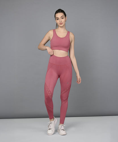 A woman wearing a Peony Pink Netted Top with a honeycomb self pattern and white sneakers stands against a plain gray background.