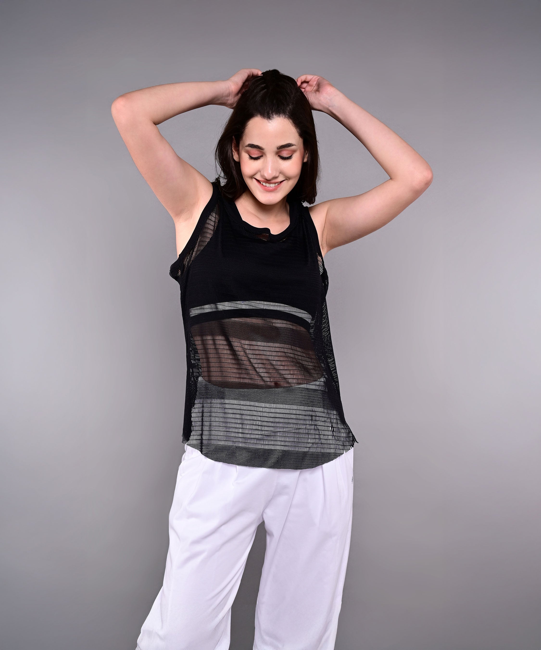 A woman smiles with hands behind her head, wearing the Tuna Active Black Netted Tank Top and white pants against a gray background.
