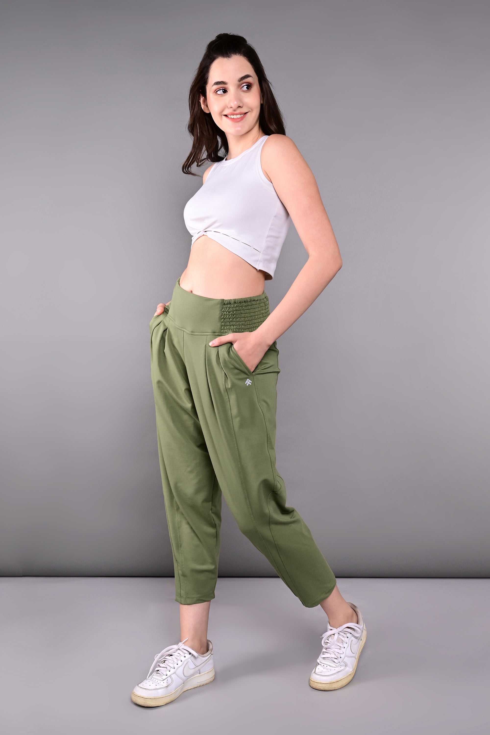 A woman smiles against a gray background wearing a White Twisted Crop Top, olive green pants, and white sneakers.