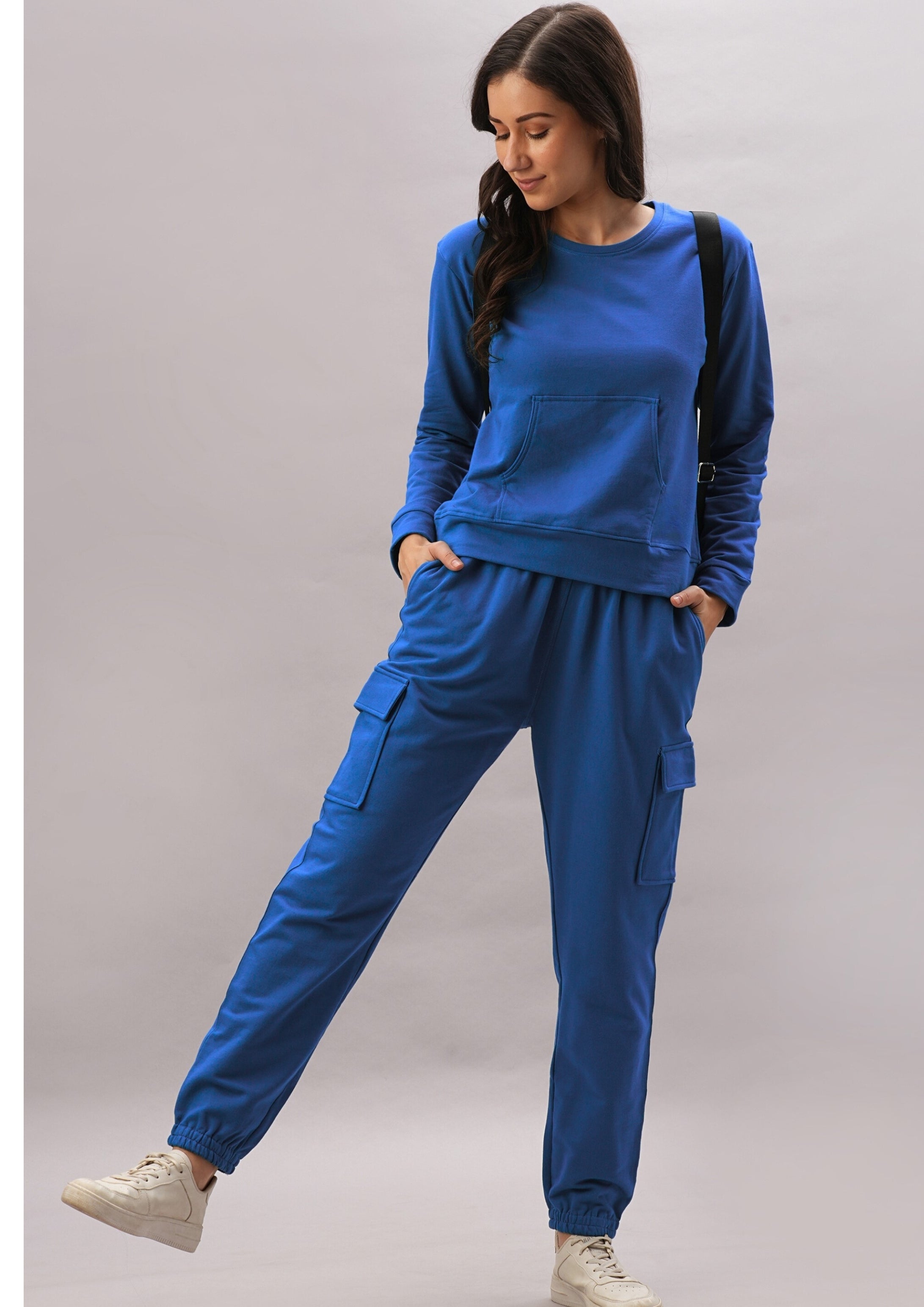 A woman wears the Soft Royal Blue Trackset, featuring cargo-style joggers made from a breathable cotton-viscose blend, paired with white sneakers and a black backpack.
