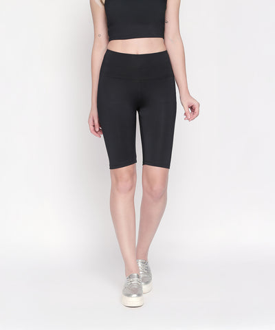 A woman stands against a white background wearing Black Biker/Cycling Shorts made from four-way stretch fabric, paired with a black crop top and silver sneakers.