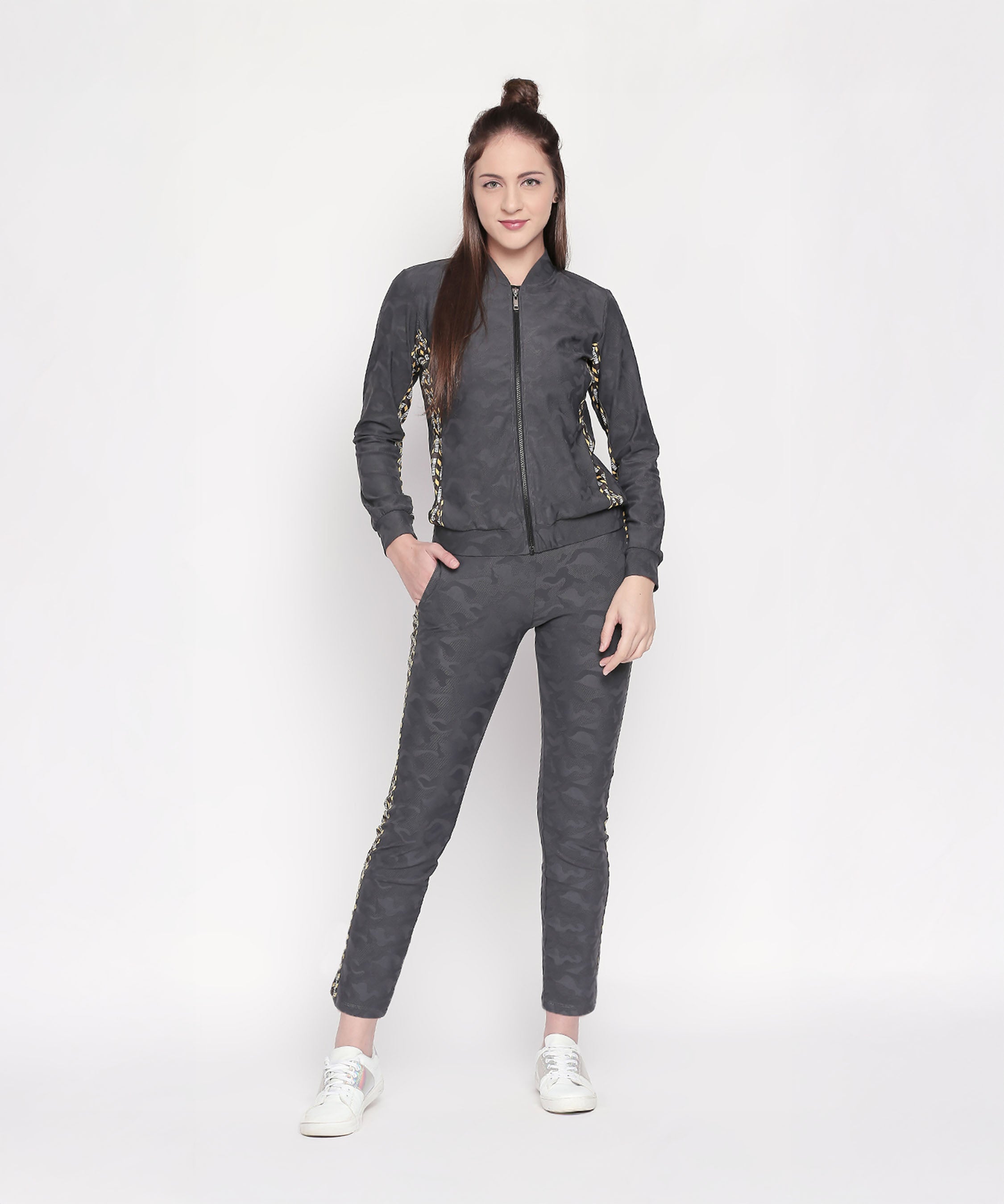 Woman in a Camouflage Print Tracksuit with dark grey elasticated track pants, gold detailing, and a V-neck collar jacket, standing and smiling against a white background.