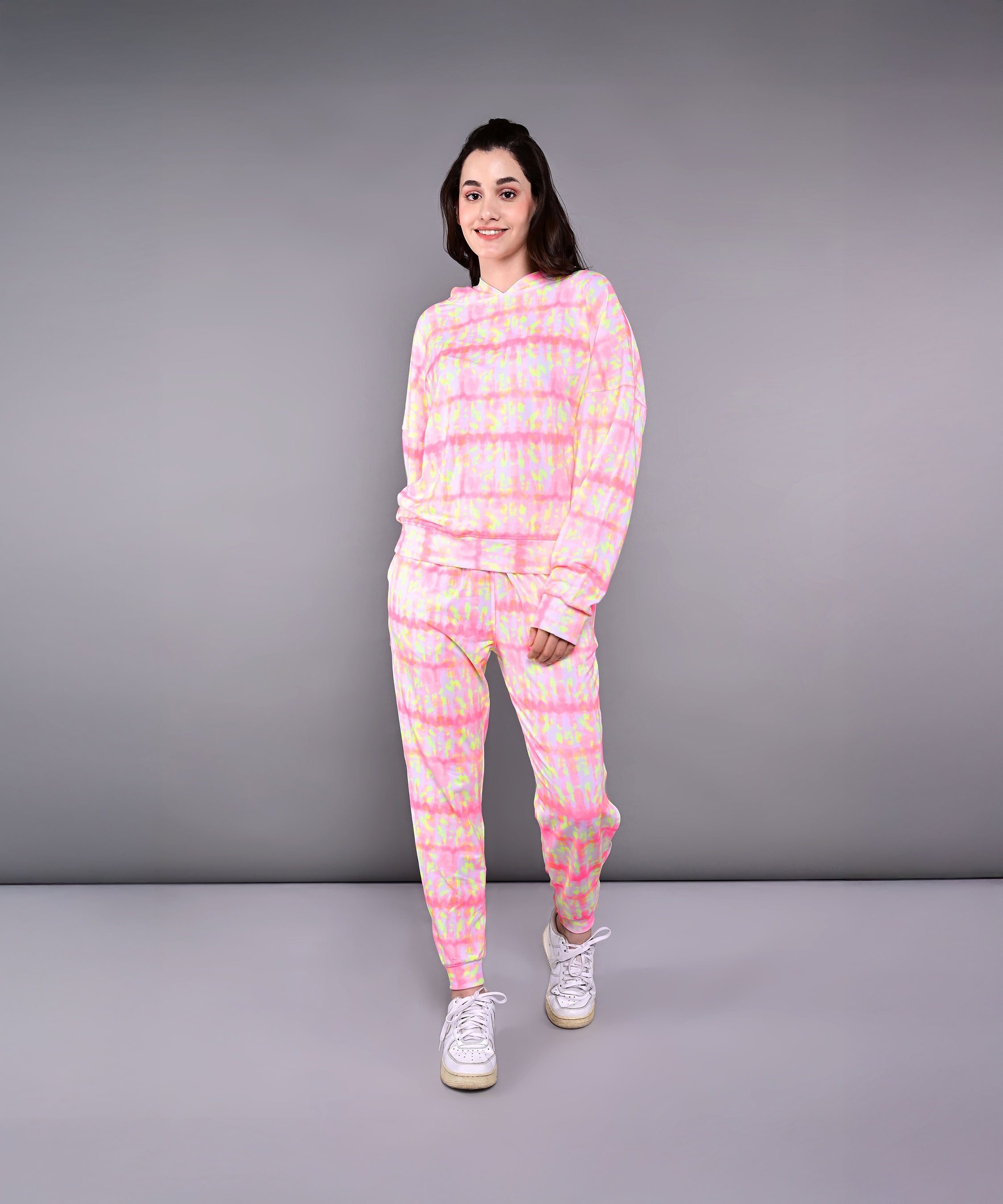 Woman in the Comfort Fit Tie-Dye Sweatshirt With Joggers Set, paired with white sneakers, stands and smiles against a gray background.