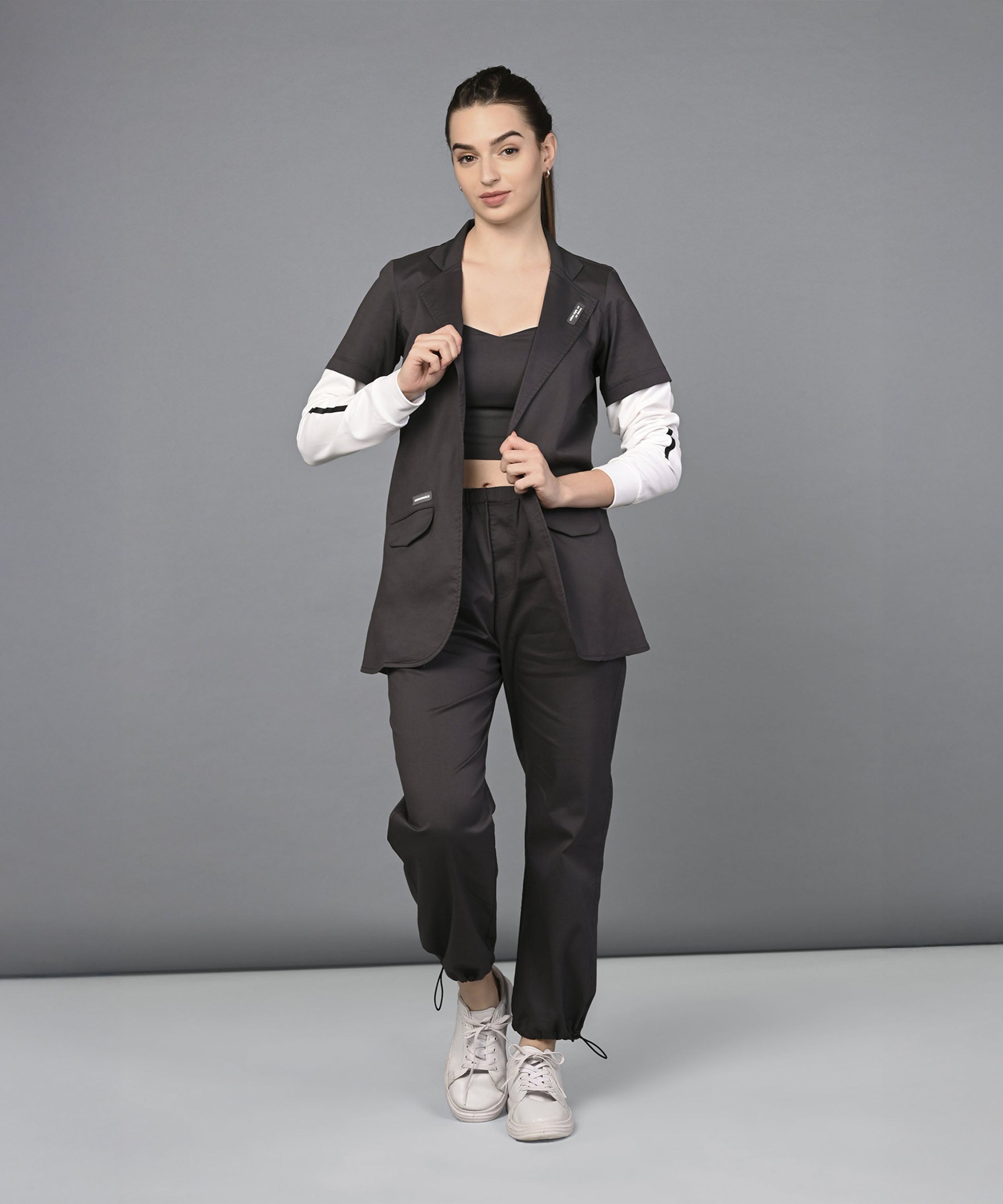 Woman wearing the Athleisure Blazer Tracks Two Piece Co-ord set, a black suit with white sleeves, paired with a black crop top and white sneakers, stands against a gray background.