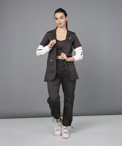 Woman wearing the Athleisure Blazer Tracks Two Piece Co-ord set, a black suit with white sleeves, paired with a black crop top and white sneakers, stands against a gray background.