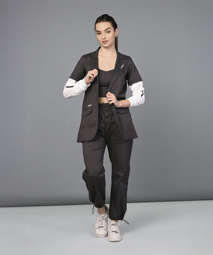 Woman wearing the Athleisure Blazer Tracks Two Piece Co-ord set, a black suit with white sleeves, paired with a black crop top and white sneakers, stands against a gray background.