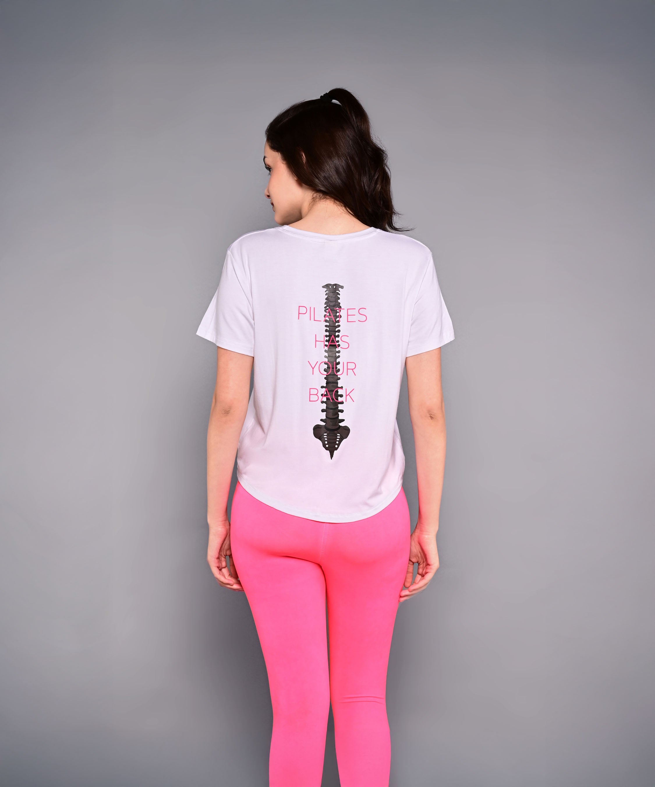 Wearing the 'Pilates Has Your Back' Printed Tee, this woman sports pink leggings and a white cotton-modal shirt with a spine graphic—an ideal motivational top for her next workout.
