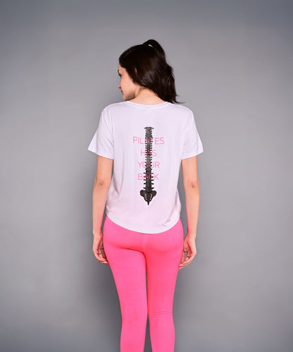Wearing the 'Pilates Has Your Back' Printed Tee, this woman sports pink leggings and a white cotton-modal shirt with a spine graphic—an ideal motivational top for her next workout.