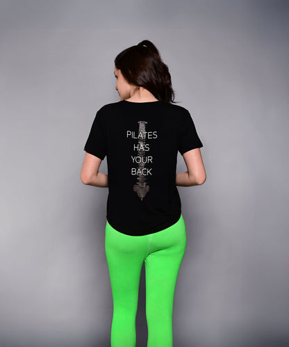 A woman in bright green leggings wears the 'Pilates Has Your Back' Printed Tee, featuring the motivational slogan on the back—perfect for workouts or casual wear.