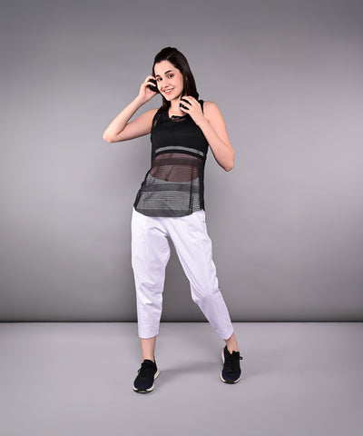 Woman smiles and poses in the Tuna Active Black Netted Tank Top with white pants against a plain gray background.