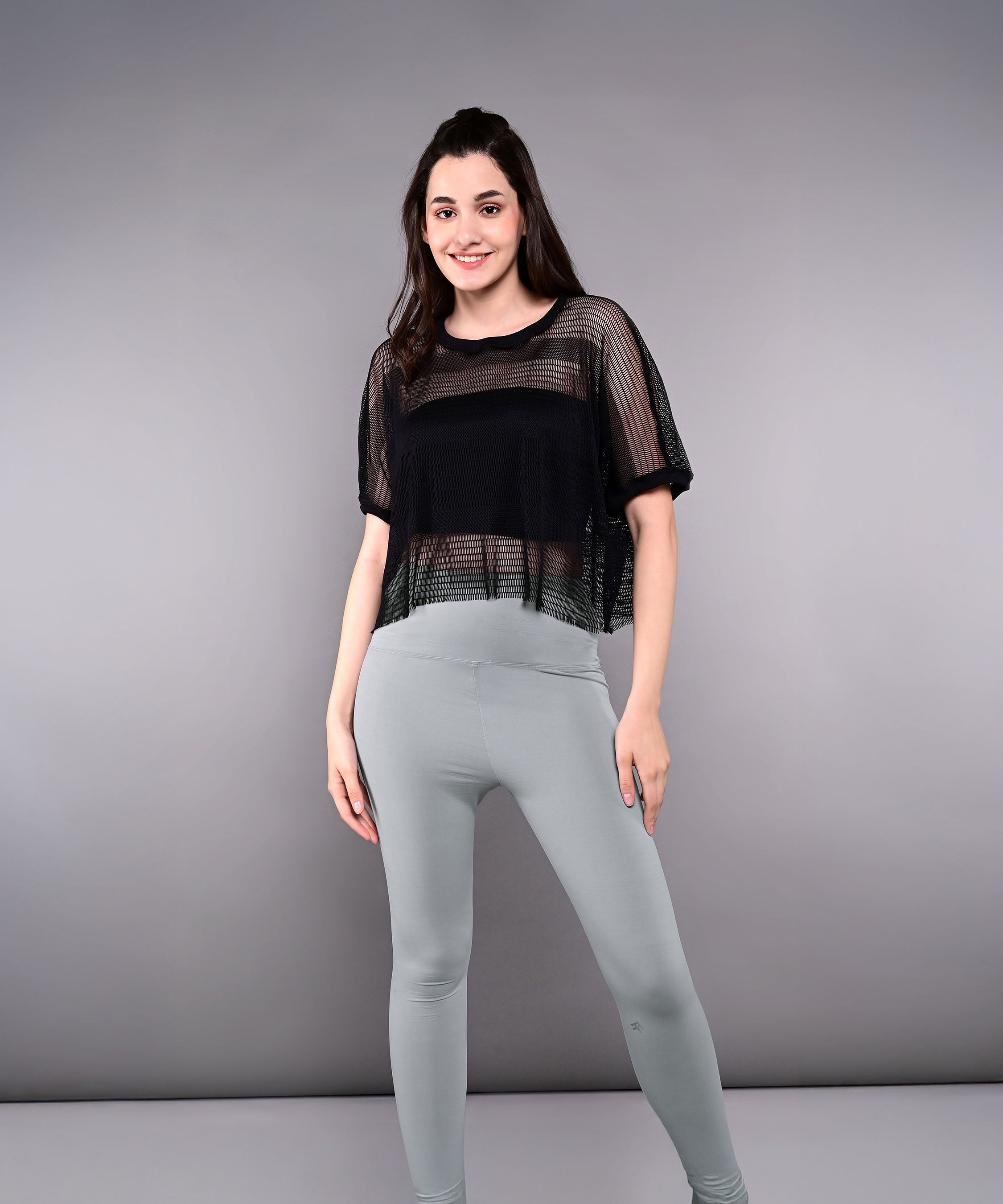 Woman smiles against a plain gray background wearing the Tuna Active Black Netted Top and light gray leggings.