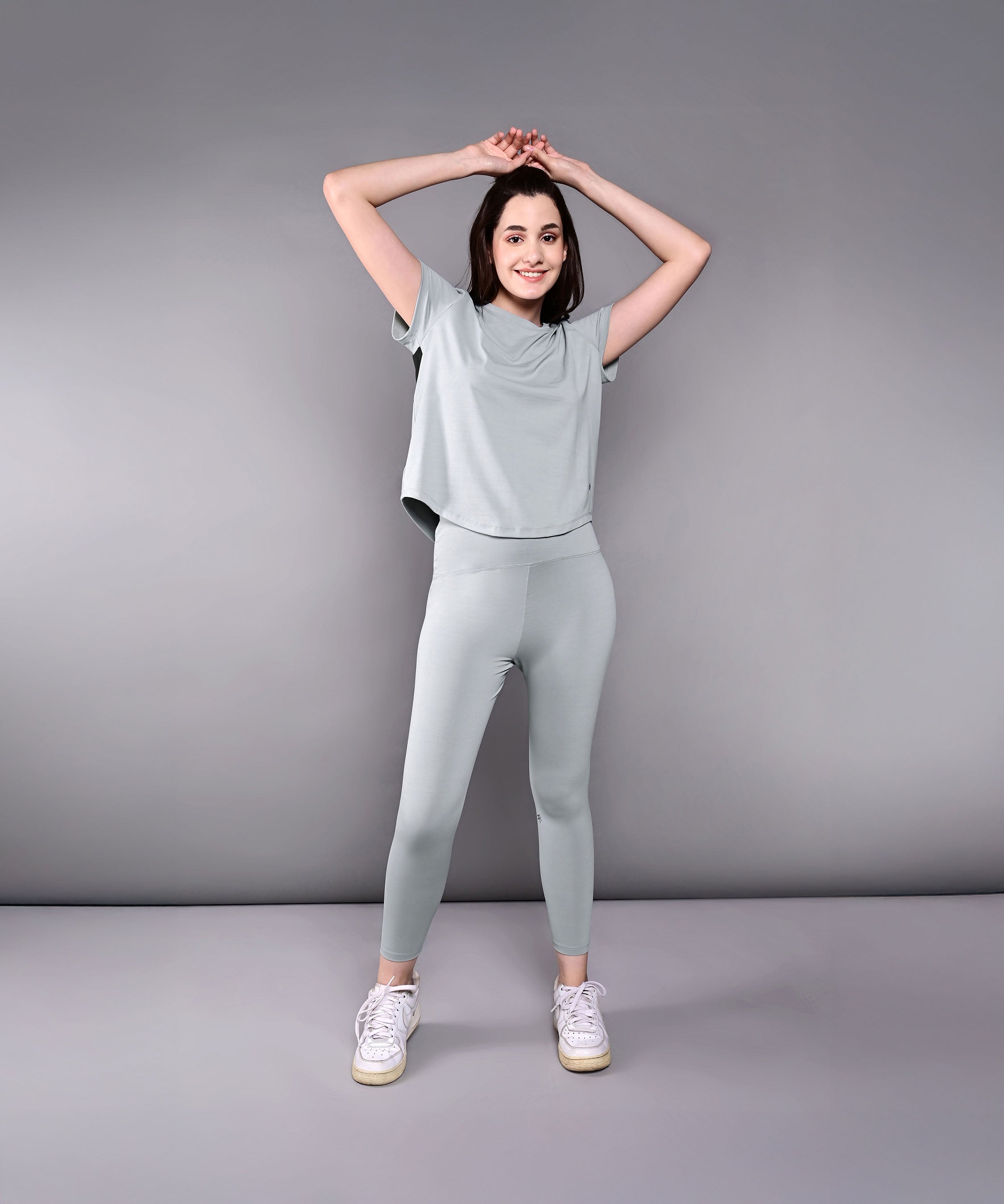 Model wears the Dynamic Grey Performance Set, featuring light green athletic leggings and a grey racerback top, posing with arms raised against a plain gray background.