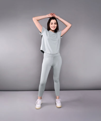 Model wears the Dynamic Grey Performance Set, featuring light green athletic leggings and a grey racerback top, posing with arms raised against a plain gray background.