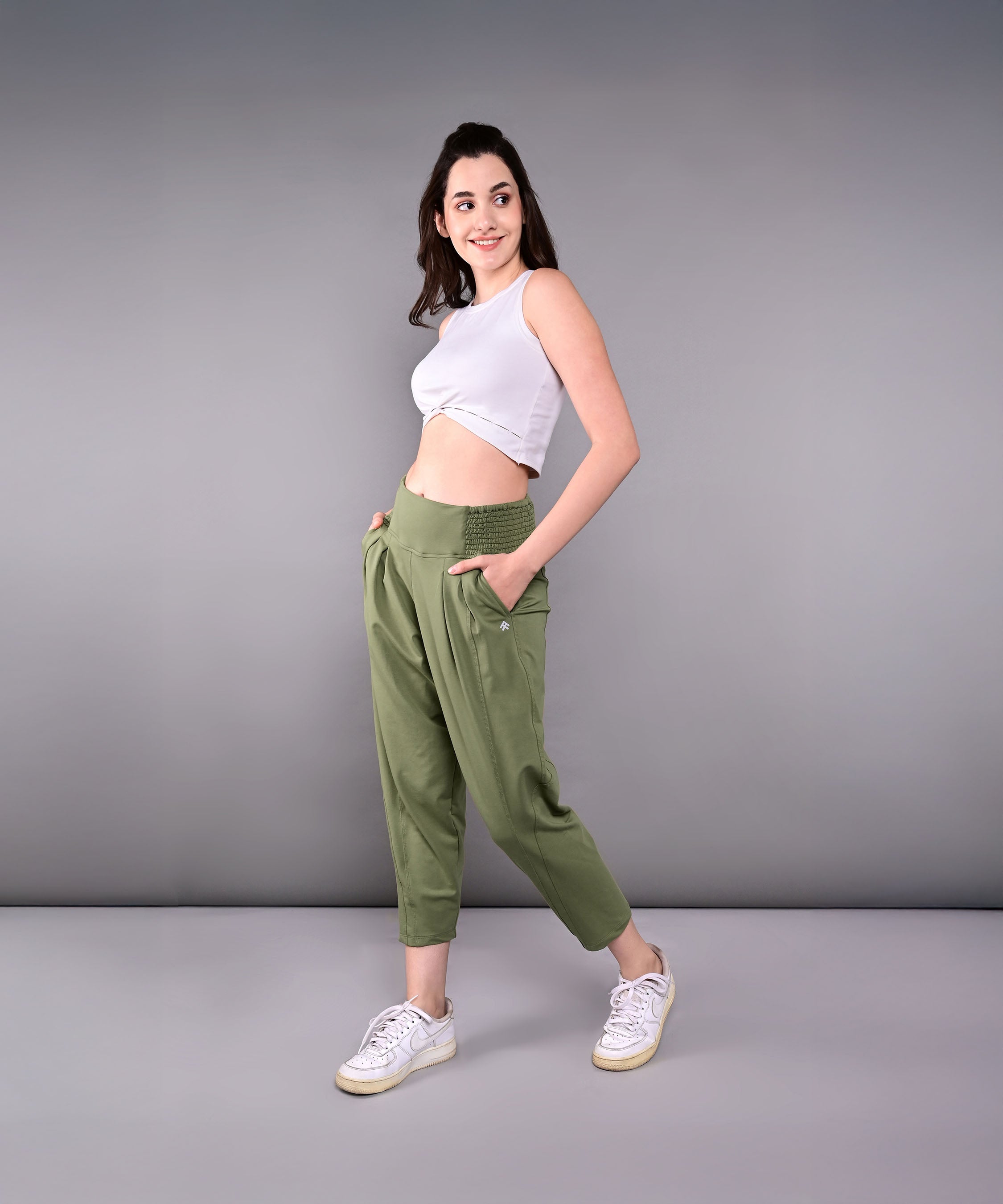 A woman in a white crop top and green Pleated Yoga Pant With Smocked Waistband stands sideways, smiling with hands in pockets, against a gray background.