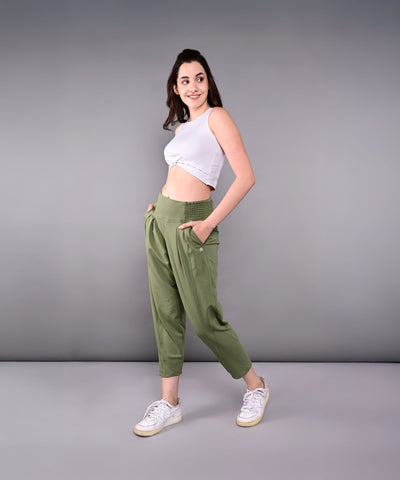 A woman in a white crop top and green Pleated Yoga Pant With Smocked Waistband stands sideways, smiling with hands in pockets, against a gray background.