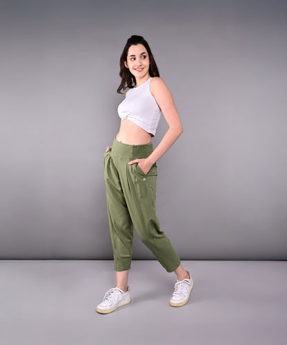 A woman in a white crop top and green Pleated Yoga Pant With Smocked Waistband stands sideways, smiling with hands in pockets, against a gray background.