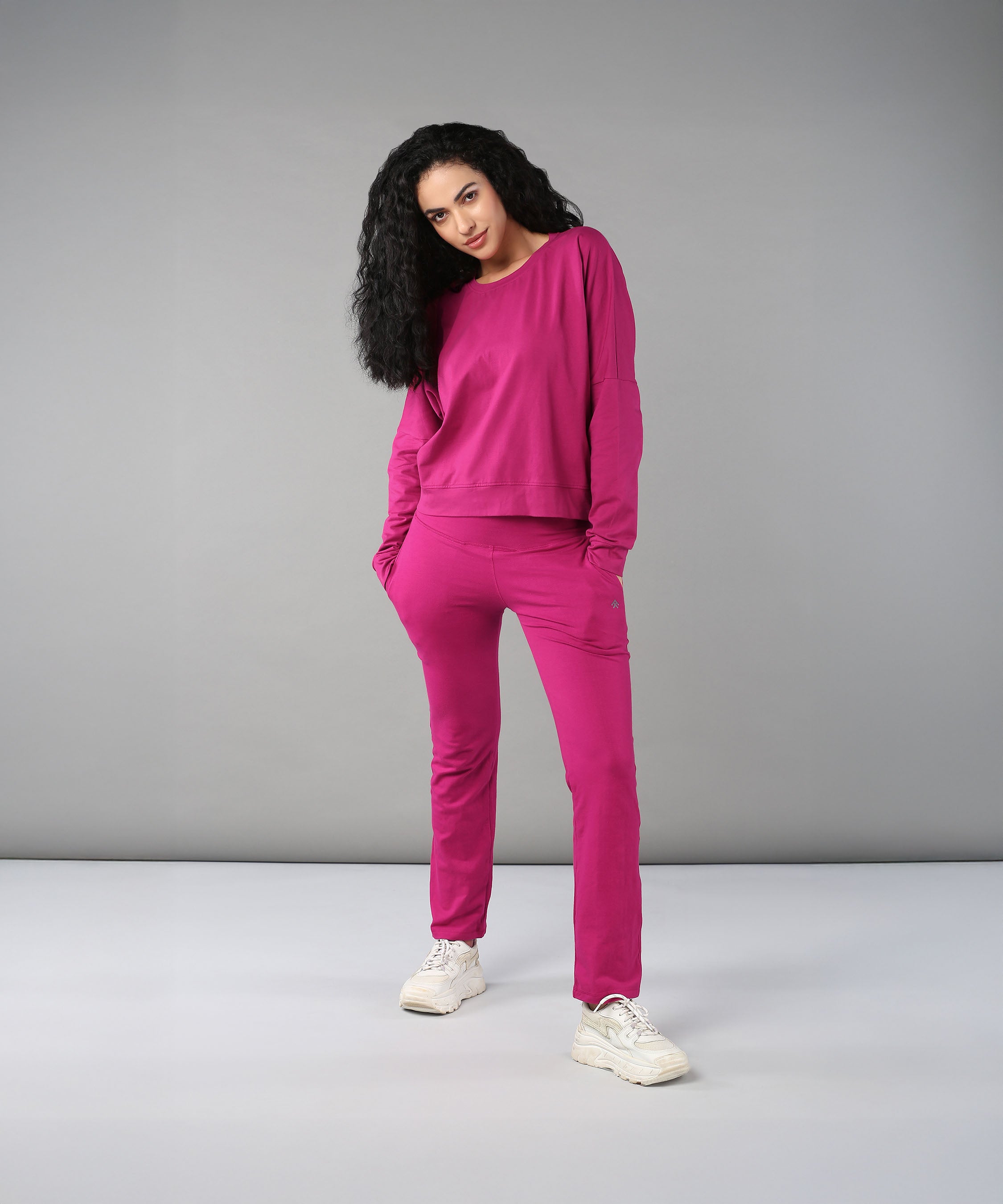 Model with curly hair wears the Deep Pink Comfort Duo—cotton lycra top and joggers—paired with white sneakers, standing against a gray background.