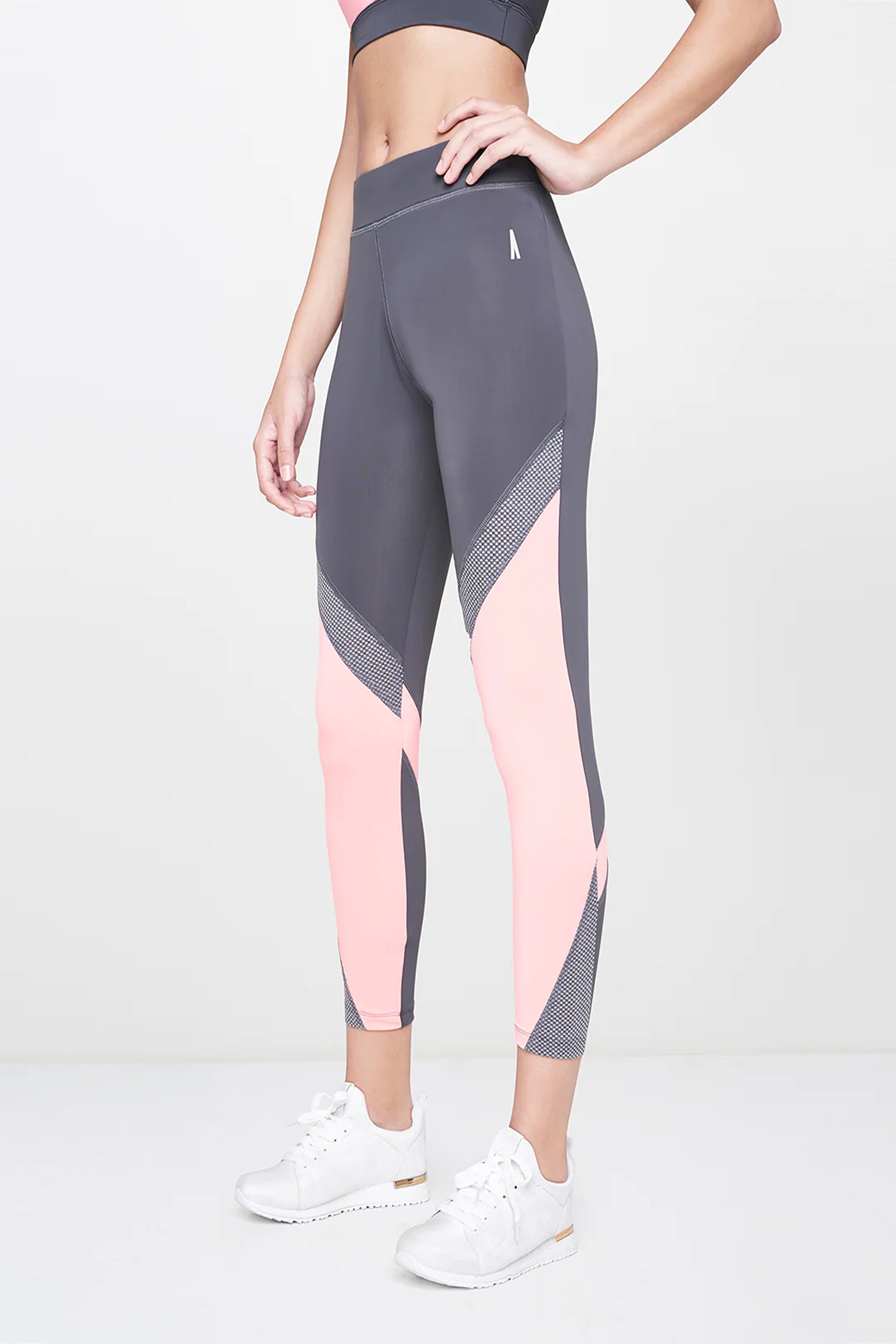 A woman wears Grey Leggings featuring pink colorblock, paired with a sports bra and white sneakers, standing against a plain background.