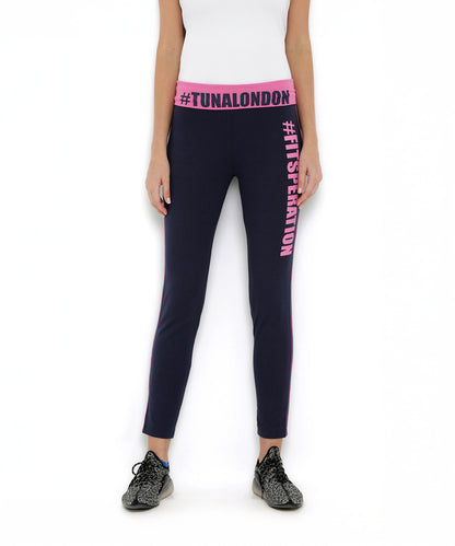 A woman stands on a white background wearing Tuna London Solid Women's Blue Track Pants, featuring a pink waistband and text, paired with black sneakers and a white top.