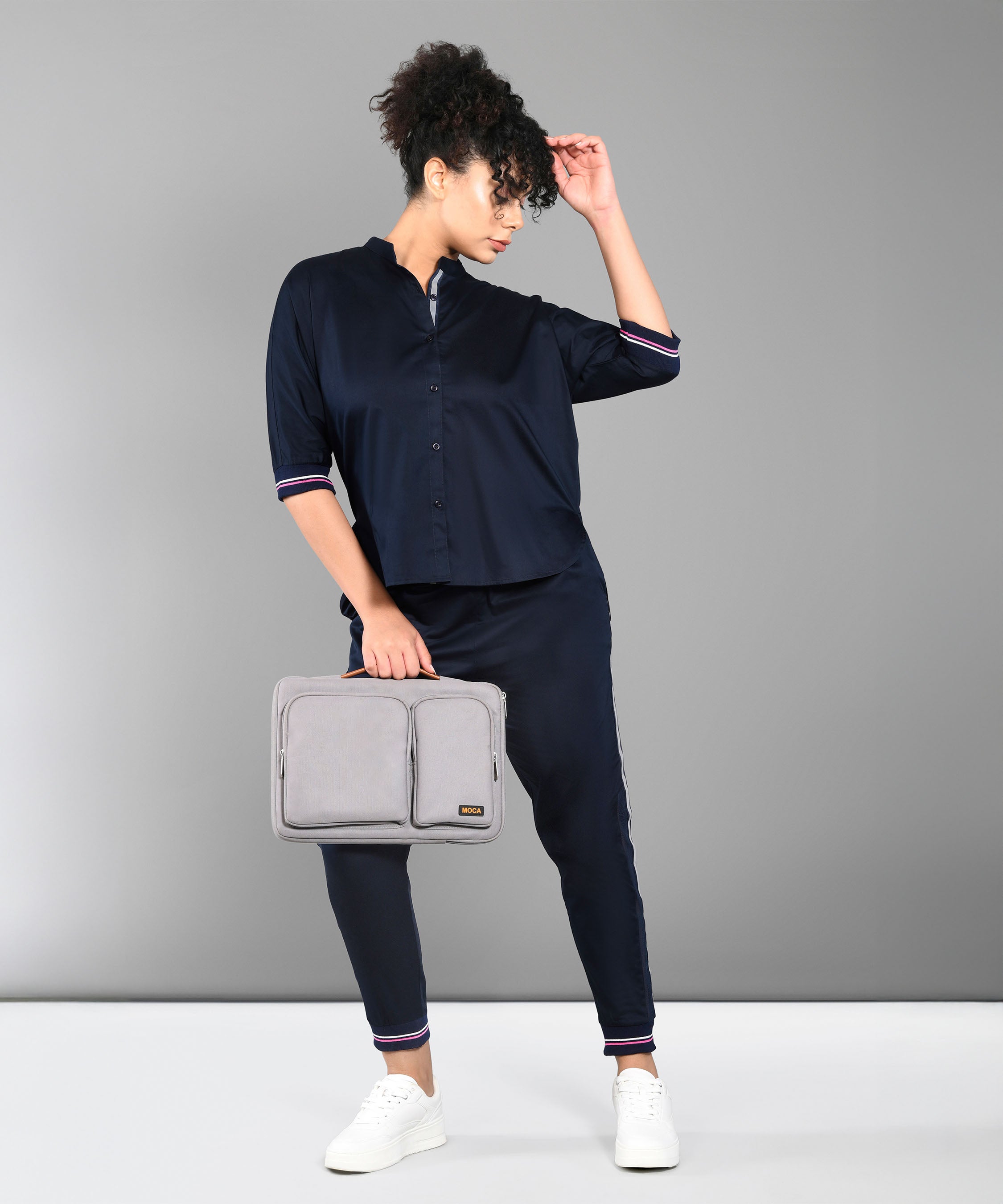 Woman in the Accented Set, a navy cotton co-ord, stands against a plain gray background holding a gray laptop bag.