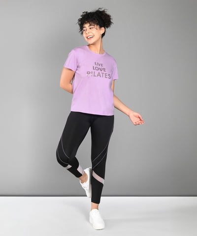 Woman in activewear smiling, wearing a Pilates Pink T-shirt, black leggings, and white sneakers.