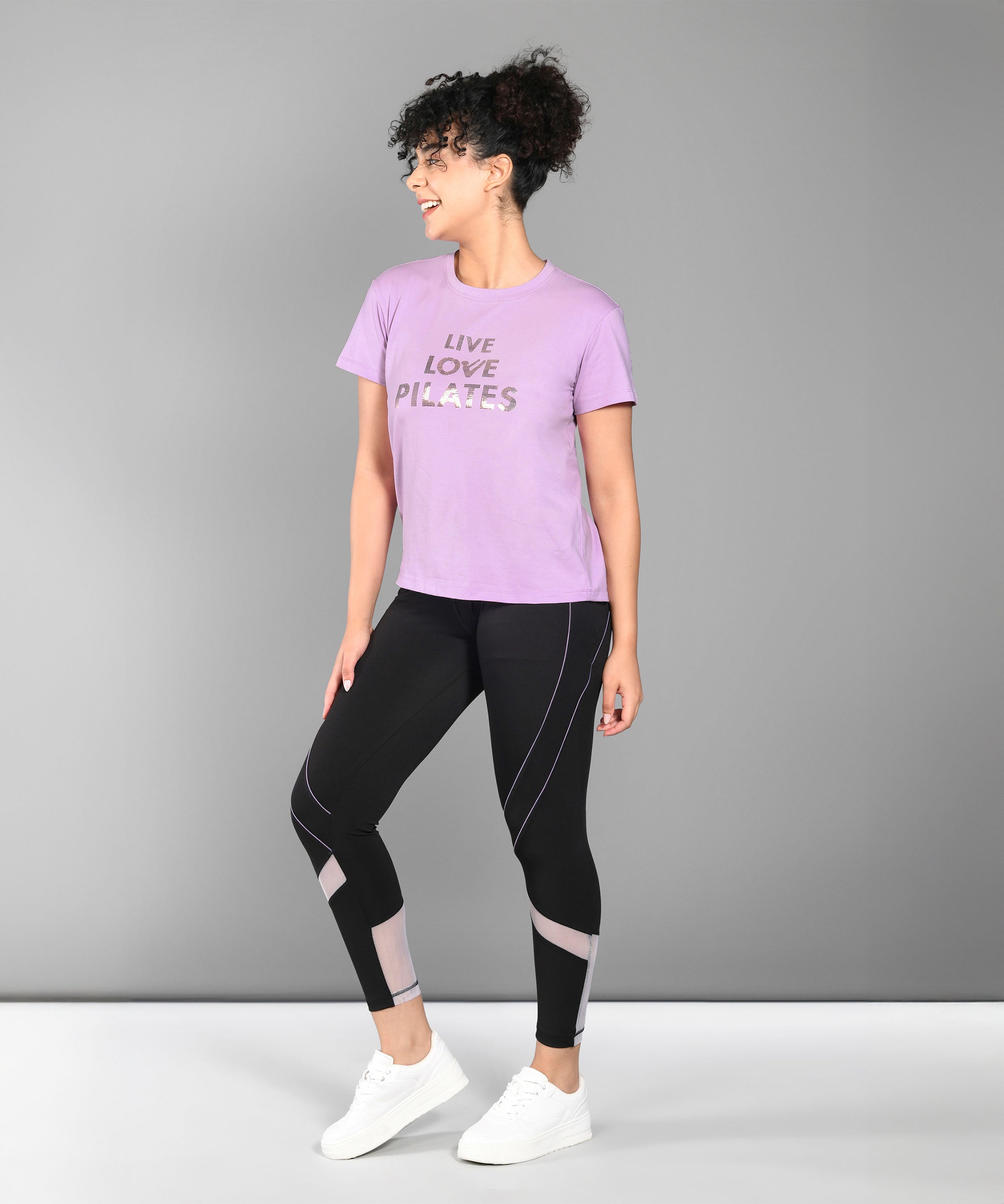 Woman in a Pilates Pink T-shirt, black leggings, and white sneakers standing and smiling.