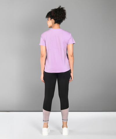A woman faces away, wearing a Pilates Pink T-shirt, black leggings with gray cuffs, and white sneakers.
