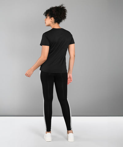 A person stands facing away, wearing a Pilates Black T-shirt, black leggings with white stripes, and white sneakers.