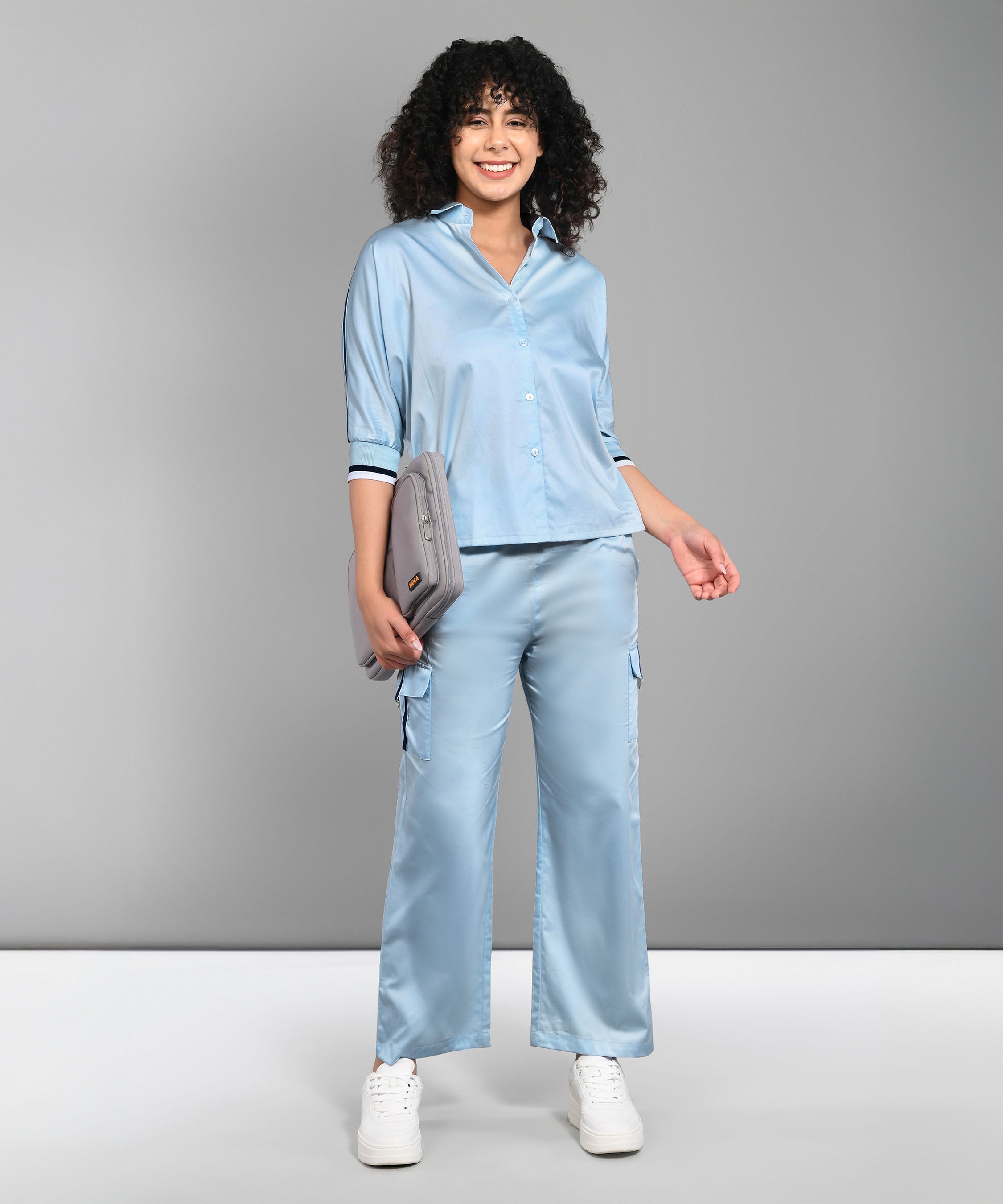 A woman smiles against a gray background, wearing the Denim Chic Set—a light blue batwing sleeve shirt and matching pants—while holding a gray clutch.