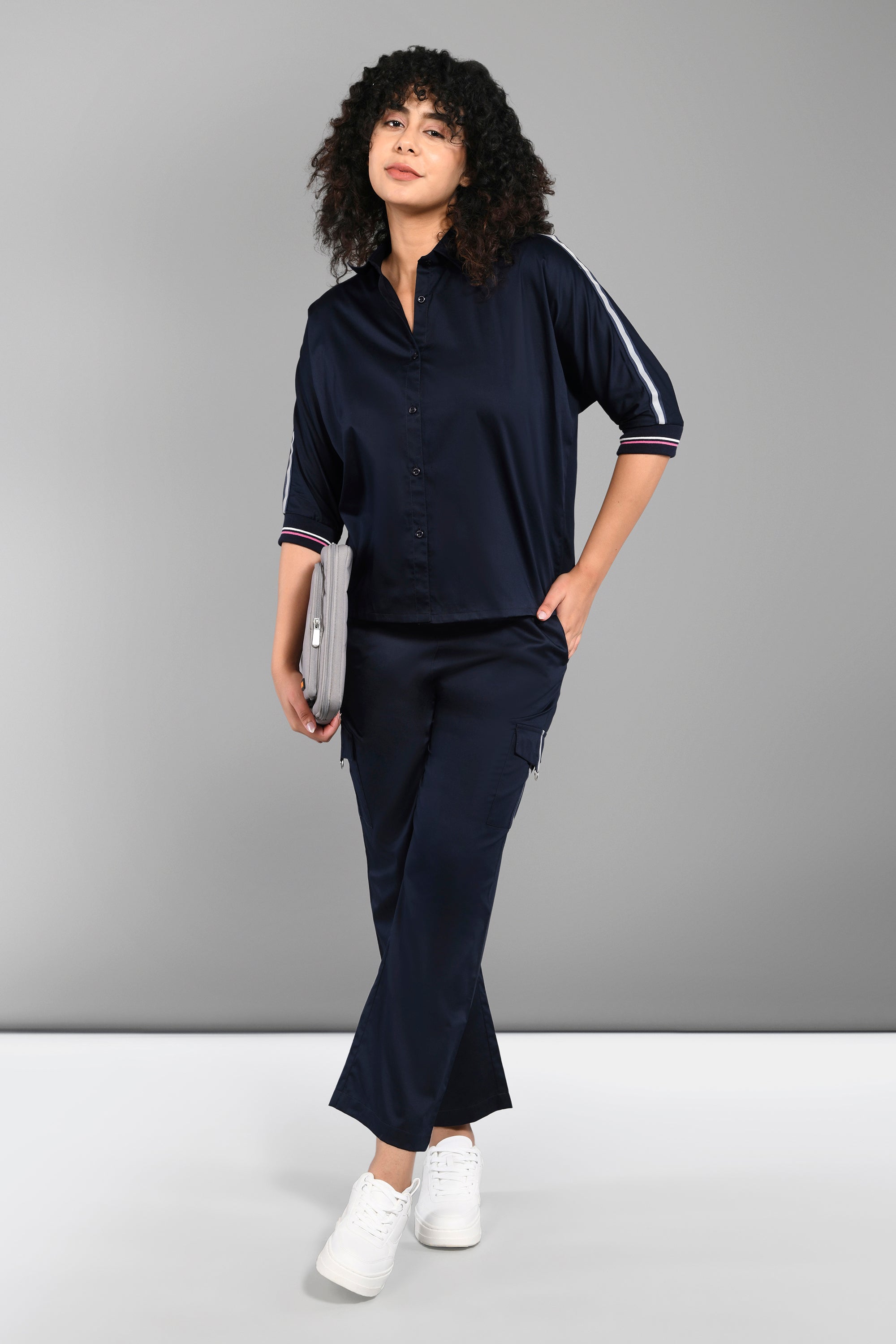 Woman in the Navy Blue Chic Set—navy co-ord with batwing sleeve shirt and cargo track pant—holds a laptop, wears white sneakers, and stands against a plain grey background.