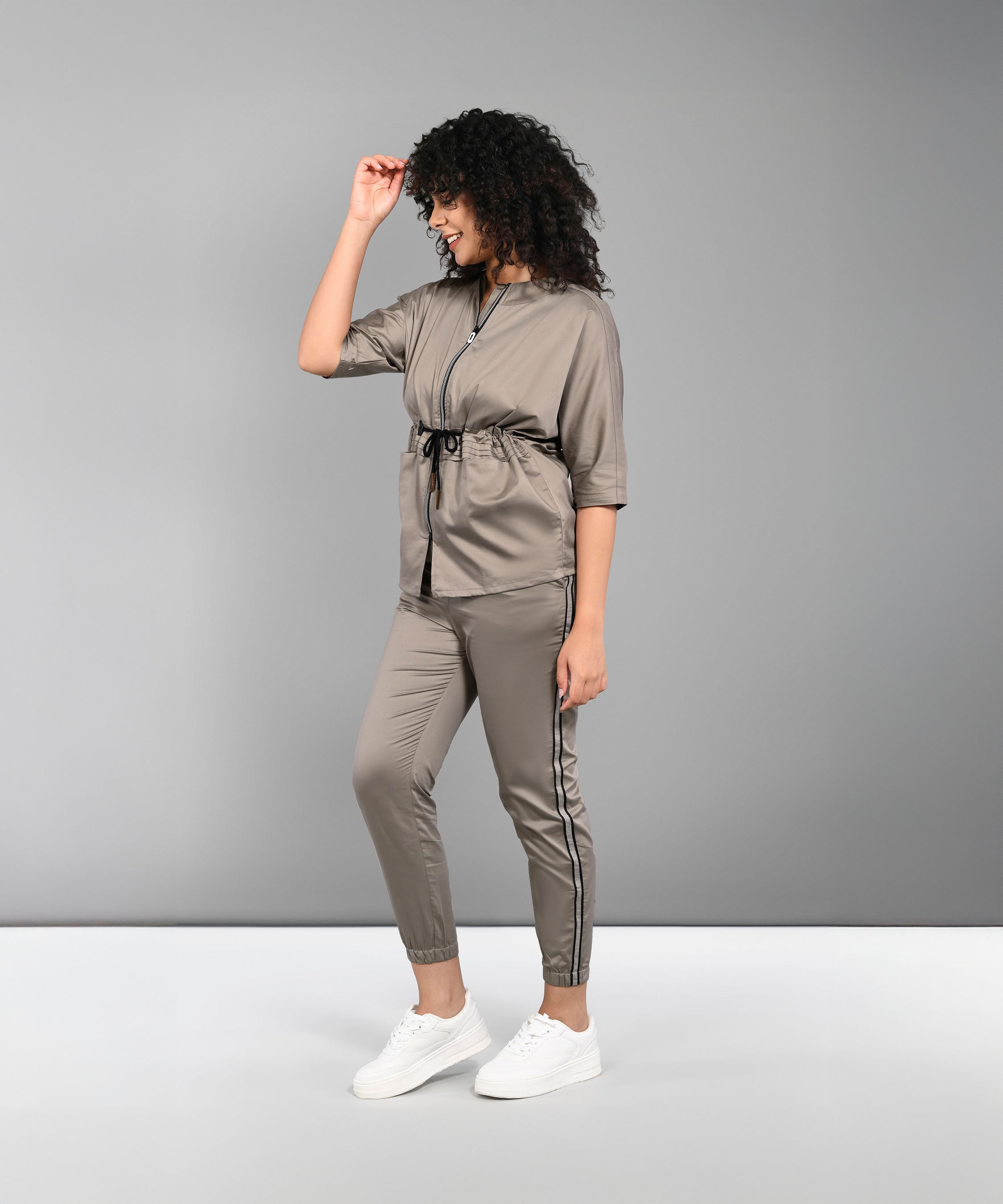 A woman models the Zipped Neutral Jacket Jogger Set—crafted from 100% cotton in taupe—with white sneakers, posing and smiling sideways against a plain gray background.