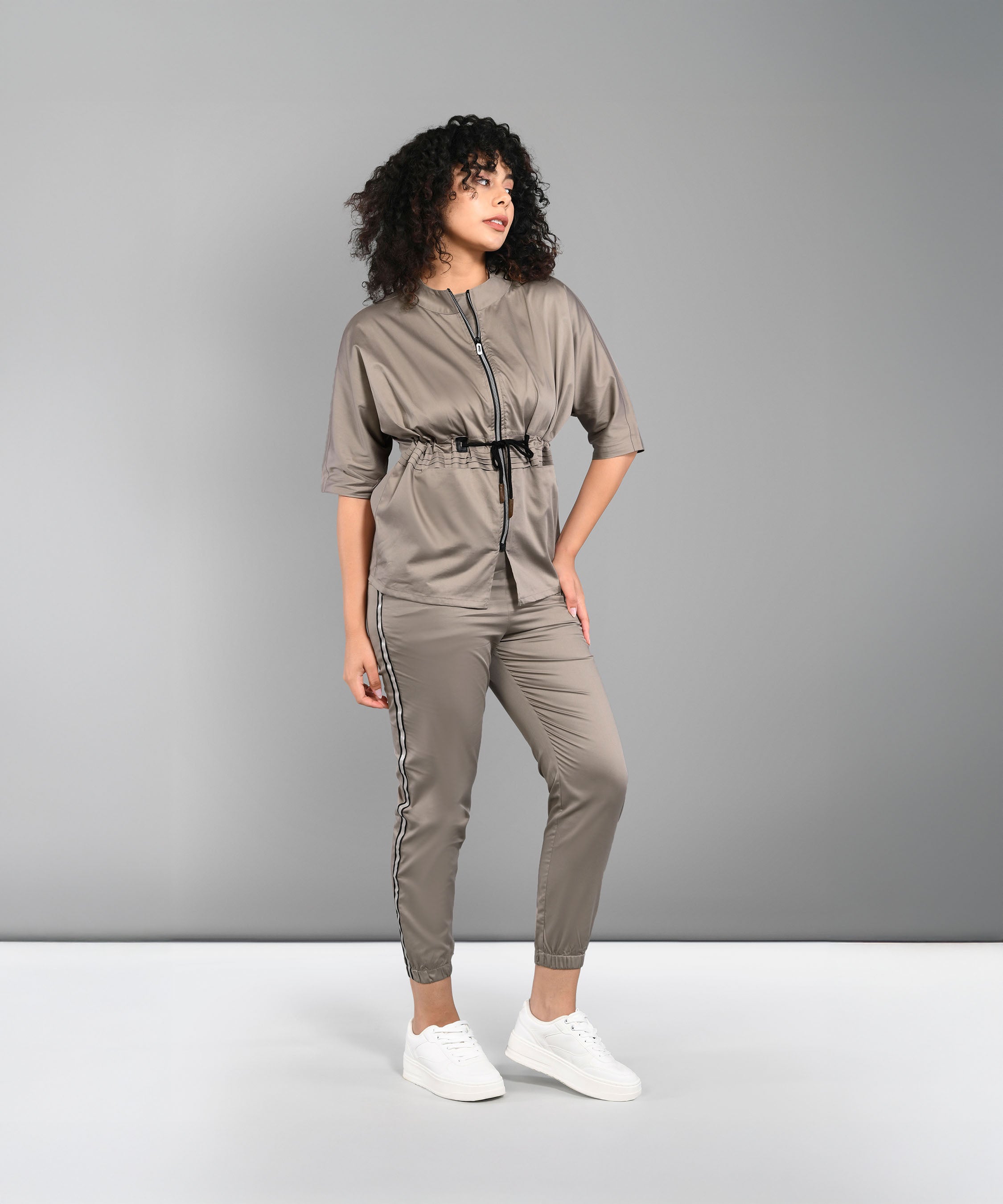 A woman in the Zipped Neutral Jacket Jogger set with a black zipper and white sneakers stands against a gray background.