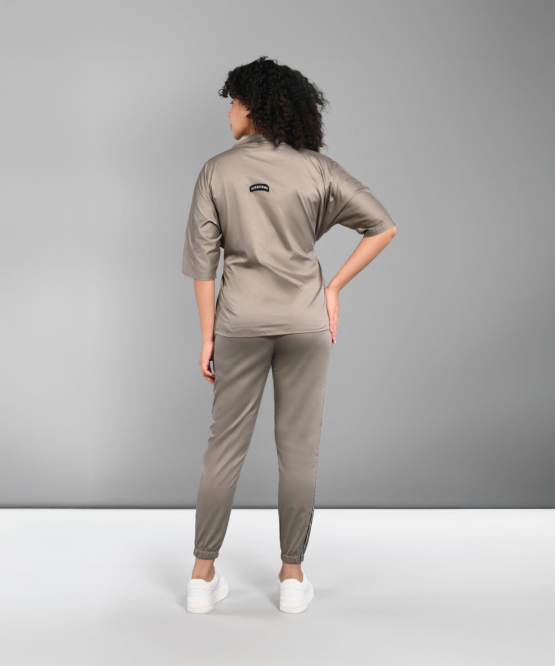 Woman with curly hair wears the Zipped Neutral Jacket Jogger set, made from 100% cotton in taupe, paired with white sneakers and standing with her back to the camera.