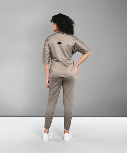 Woman with curly hair wears the Zipped Neutral Jacket Jogger set, made from 100% cotton in taupe, paired with white sneakers and standing with her back to the camera.