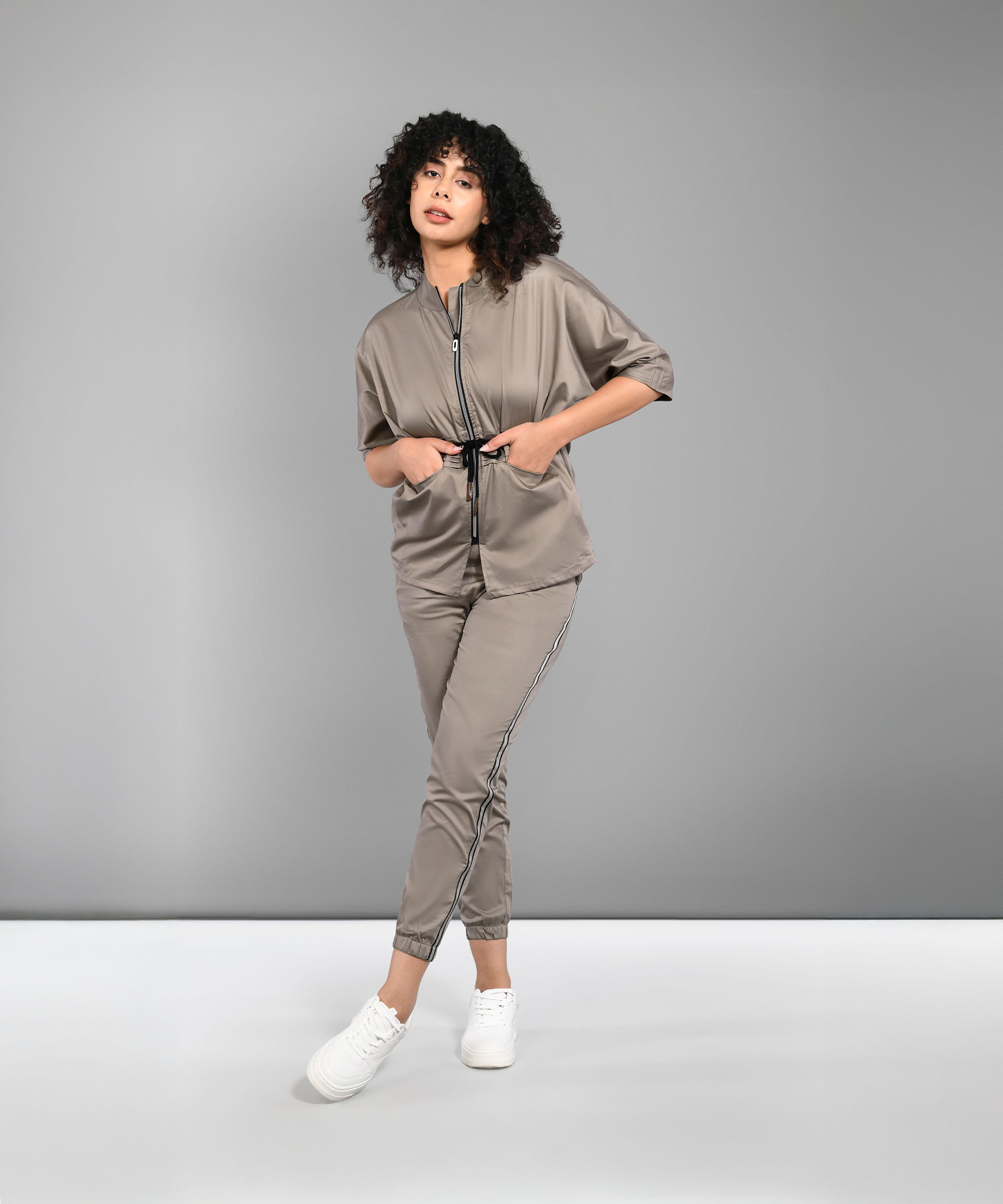 A woman models the Zipped Neutral Jacket Jogger set—100% cotton taupe joggers and a zip-up top with drawstring—paired with white sneakers, standing against a plain gray background.