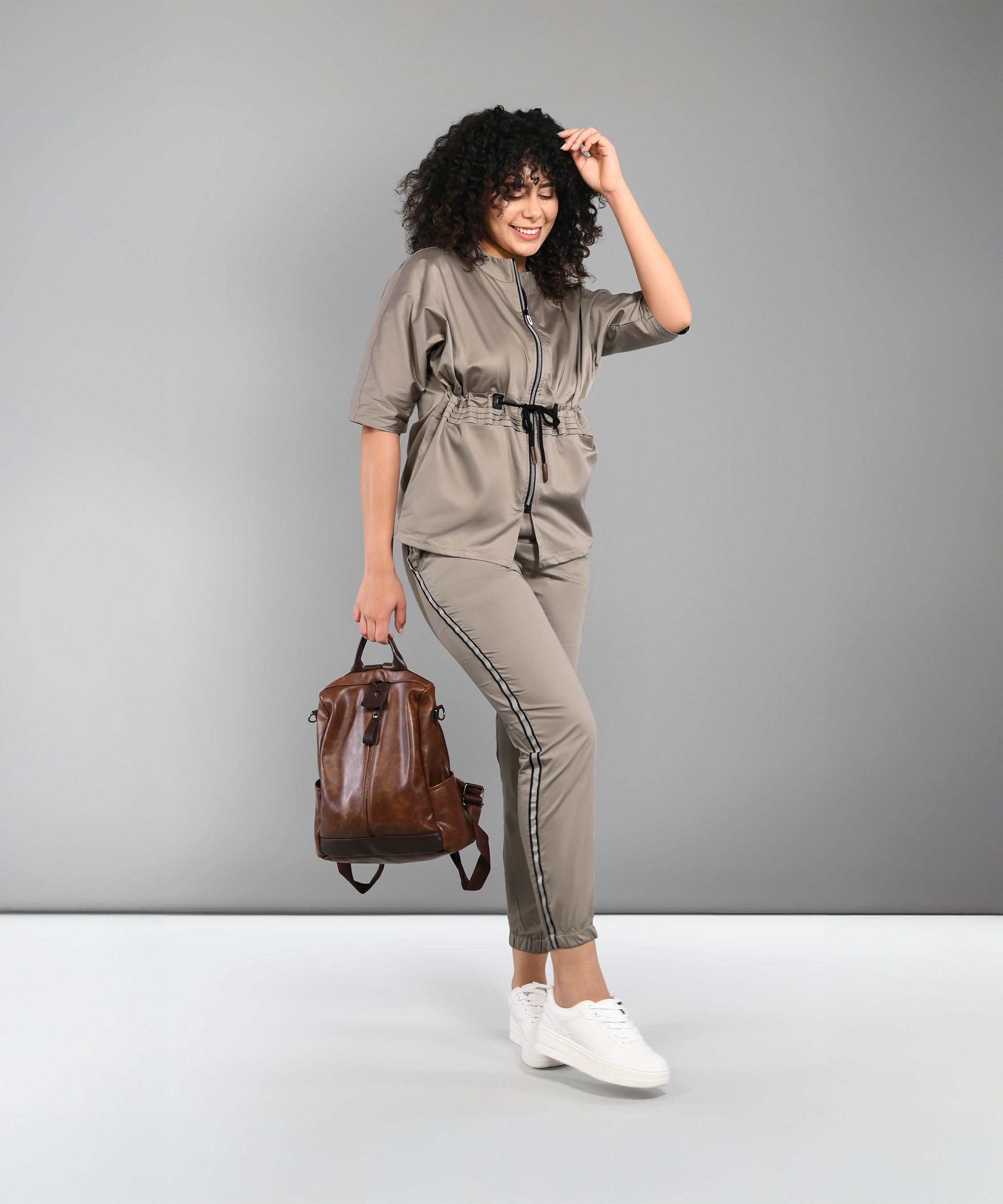 Woman wearing the Zipped Neutral Jacket Jogger Set made from 100% cotton, holding a brown backpack and smiling against a gray background.