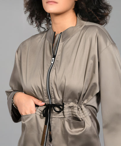 Woman wearing the Zipped Neutral Jacket Jogger Set in shiny taupe 100% cotton, featuring a zip-up jacket, drawstring waist joggers, and pockets, posed against a gray background.